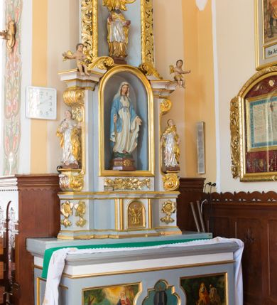 Zdjęcie nr 2: Ołtarz architektoniczny, dwukondygnacyjny, jednoosiowy ze zwieńczeniem. Ołtarz prostopadłościenny z drewnianą mensą i kamiennym portatylem. Antependium ozdobione trzema płycinami z czego środkowa w kształcie czwórliścia przenikającego się z kwadratem; dwie boczne, ustawione antytetycznie, w kształcie leżącego prostokąta z parą boków wewnętrznych w kształcie łuku nadwieszonego wklęsłego. W płycinie środkowej polichromowany monogram maryjny, w płycinach bocznych po lewej stronie Zwiastowanie, a po prawej Ucieczka do Egiptu. 

Nastawa na niewysokim, uskokowym cokole o ukośnie ustawionych bokach, zdobionych słonecznikami. W środkowej partii cokołu wkomponowane tabernakulum w formie prostopadłościennej szafki na profilowanym, uskokowym cokole z narożami ujętymi dwoma parami pilastrów z półwałkiem pośrodku trzonu, wspierających profilowany, uskokowy gzyms. Drzwiczki tabernakulum w kształcie prostokąta zamkniętego łukiem półkolistym, w polu płaskorzeźbiony kielich z Hostią oraz kłosami zbóż. Kondygnacja flankowana parą zdwojonych pilastrów oraz dostawionych, ustawionych pod kątem pilastrów z nałożonymi spływami wolutowymi. Podpory wspierają przerywane, niepełne belkowanie oraz przyczółek. Pole środkowe zajmuje wnęka w kształcie stojącego prostokąta, zamkniętego półkoliście. W polu figura Matki Boskiej Niepokalanie Poczętej. Pod wnęką podwieszona, rzeźbiona girlanda. Po lewej stronie kondygnacji, na cokole ułożonym na zakończeniu woluty ustawiona jest rzeźba św. Jana Ewangelisty, a po drugiej stronie figura nierozpoznanej świętej. Na belkowaniu usadzona jest para aniołków. Druga kondygnacja w formie wnęki w kształcie stojącego prostokąta, zamkniętego półkoliście, flankowana po bokach filarami, ozdobionymi liśćmi akantu. W polu umieszczona rzeźba św. Mikołaja. Nad rzeźbą stylizowana duża muszla palmetowa z podwieszonymi liśćmi akantu. Zwieńczenie w formie promienistej glorii z Okiem Opatrzności pośrodku, ujętej wieńcem srebrzonych obłoków. Struktura polichromowana na biało, detal architektoniczny i ornamentyka złocone oraz srebrzone. Na cokole tabliczka z napisem: „FUNDACJA / SREBRZENIE ŚWIECZNIKÓW / OŁTARZA NMP / Franciszek i Małgorzata Łojek / 2003”. 