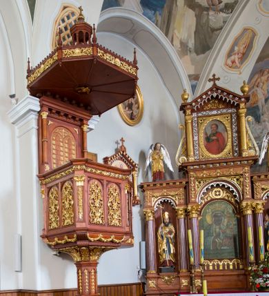 Zdjęcie nr 1: Przyścienną ambonę wspartą na masywnym filarze ustawiono przy północnej ścianie nawy, obok wejścia do kaplicy Matki Boskiej Szaflarskiej. Wejście na nią prowadzi z kruchty północnej. Ambona z wejściem połączona jest za pomocą podestu z drewnianą, ażurową balustradą, utworzoną z półokrągłych arkadek wspartych na wysmuklonych tralkach. Filar ambony ustawiono na dwupoziomowym cokole na planie czworoboku o ściętych narożach. Cokół w górnej części przechodzi w ośmioboczny filar zwieńczony stylizowanymi i złoconymi liśćmi akantu ułożonymi kielichowo. Boki filaru zdobią stylizowane, złocone kampanulle. Kosz ambony wykonano na planie czworoboku. W narożach pojawiły się pilastry podtrzymujące wyłamane belkowanie stanowiące parapet ambony, dekorowane stylizowaną wicią akantową z palmetami. Ścianki kosza ozdobione zostały podwójnymi, prostokątnymi płycinami, zamkniętymi łukiem odcinkowym, które wypełniono płaskorzeźbioną i złoconą, stylizowaną wicią akantu. Również pilastry na narożach ozdobiono stylizowaną wicią liści akantu, wieńcząc je kapitelami kompozytowymi. Przy dolnej krawędzi kosza zawieszono płaskorzeźbione i złocone girlandy kwiatowo-roślinne. Wysoki zaplecek ambony uzyskał architektoniczną formę flankowaną przez dwie kolumny z attyckimi bazami na wysokich postumentach, z pierścieniem w połowie wysokości i kapitelami kompozytowymi. Kolumny podtrzymują belkowanie dodatkowo wsparte na czterech konsolkach. W polu pomiędzy kolumnami na tle prostokątnej zwieńczonej półokrągło płyciny wyobrażono dwie płaskorzeźbione, półokrągłe tablice Dekalogu, dekorowane diagonalnymi rozetkami. Ponad amboną zawieszono baldachim na planie czworoboku o ściętych narożach, zwieńczonych małymi pinaklami. Jego boki ozdabia stylizowany ornament akantowy z muszlami. Na podniebieniu wyobrażono rzeźbioną gołębicę Ducha Świętego. Całość baldachimu wieńczy ażurowa latarnia na planie ośmioboku, o ośmiu kolumnach wspierających belkowanie, z daszkiem zwieńczonym krzyżykiem.