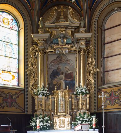 Zdjęcie nr 9: Nastawa ołtarzowa jednoosiowa, jednokondygnacyjna ze zwieńczeniem, ustawiona na wysokim dwustrefowym cokole, zryzalitowanym na zewnetrznych krawędziach. Ołtarz prostopadłościenny z kamienną mensą. Antepedium drewniane, dekorowane na wybrzuszeniu trzema płycinami z rytą wicią roślinną i monogramem IHS. Tabernakulum w kształcie kapliczki na cokole w formie prostopadłościanu; ścianka frontowa ujęta półfilarami dźwigającymi gzyms opasujący go z trzech stron, na froncie przyjmujący kształt łuku segmentowego, spiętego ustawionymi przeciwstawnie wolutami. Trzony półfilarów dekorowane owocowo-roślinnymi zwisami, pole środkowe skręconymi liśćmi akantu w układzie lustrzanym. Na nim nastawa w formie półokrągłej ścianki dekorowanej płyciną w kształcie stojącego prostokąta zamkniętego łukiem pełnym zdobionym kielichem z unoszącą się ponad nim hostią w otoczeniu promieni. Drzwiczki ujęte skręconymi kolumnami dźwigającymi pełne belkowanie wyłamane na ich osiach i wyniesione w łuk na polem centralnym. Na środku fryzu uskrzydlona główka anielska. Na wyłamanych częściach belkowania wazony płomieniste. Nastawa ołtarzowa ujęta parą kolumn o kapitelach kompozytowych dźwigających fragmenty belkowania o silnie wysuniętym gzymsie, połączone architrawem przyjmującym nad polem środkowym kształt łuku pełnego. Pole środkowe niemal w całości wypełnia nisza ujęta ramą w formie stojącego prostokąta wyłamanego prostokątnie w partii górnego boku, wypełniona obrazem św. Rodziny wymiennie z wizerunkiem Matki Boskiej Niepokalanie Poczętej. Zwieńczenie na zwężającej się łukowato podstawie w formie przyczółka przerwanego pośrodku cokołem z krzyżem. Cokół w górnej strefie dekorowany płycinami, w partii ryzalitów wypełnionymi akantem mięsistym i uskrzydloną główką anielską na froncie. Rama obrazu w polu środkowym dekorowana wstęgą i liśćmi; w prostokątnym polu nad obrazem płaskorzeźbione obłoki z rzeźbą Gołębicy Ducha Świętego w glorii; powyżej w polu łuku półpostać Boga Ojca z rozłożonymi na boki rękami. Kondygnacja ujęta uszakami utworzonymi z silnie skręcanego mięsistego akantu, zwiniętego w woluty oraz guzy. Krawędzie podstawy zwieńczenia oraz gzyms przyczółka dekorowane liśćmi akantu. Struktura polichromowana w kolorze kremowym; ornamentyka, profile i kolumny złocone i srebrzone.