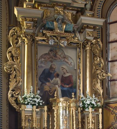 Zdjęcie nr 11: Nastawa ołtarzowa jednoosiowa, jednokondygnacyjna ze zwieńczeniem, ustawiona na wysokim dwustrefowym cokole, zryzalitowanym na zewnetrznych krawędziach. Ołtarz prostopadłościenny z kamienną mensą. Antepedium drewniane, dekorowane na wybrzuszeniu trzema płycinami z rytą wicią roślinną i monogramem IHS. Tabernakulum w kształcie kapliczki na cokole w formie prostopadłościanu; ścianka frontowa ujęta półfilarami dźwigającymi gzyms opasujący go z trzech stron, na froncie przyjmujący kształt łuku segmentowego, spiętego ustawionymi przeciwstawnie wolutami. Trzony półfilarów dekorowane owocowo-roślinnymi zwisami, pole środkowe skręconymi liśćmi akantu w układzie lustrzanym. Na nim nastawa w formie półokrągłej ścianki dekorowanej płyciną w kształcie stojącego prostokąta zamkniętego łukiem pełnym zdobionym kielichem z unoszącą się ponad nim hostią w otoczeniu promieni. Drzwiczki ujęte skręconymi kolumnami dźwigającymi pełne belkowanie wyłamane na ich osiach i wyniesione w łuk na polem centralnym. Na środku fryzu uskrzydlona główka anielska. Na wyłamanych częściach belkowania wazony płomieniste. Nastawa ołtarzowa ujęta parą kolumn o kapitelach kompozytowych dźwigających fragmenty belkowania o silnie wysuniętym gzymsie, połączone architrawem przyjmującym nad polem środkowym kształt łuku pełnego. Pole środkowe niemal w całości wypełnia nisza ujęta ramą w formie stojącego prostokąta wyłamanego prostokątnie w partii górnego boku, wypełniona obrazem św. Rodziny wymiennie z wizerunkiem Matki Boskiej Niepokalanie Poczętej. Zwieńczenie na zwężającej się łukowato podstawie w formie przyczółka przerwanego pośrodku cokołem z krzyżem. Cokół w górnej strefie dekorowany płycinami, w partii ryzalitów wypełnionymi akantem mięsistym i uskrzydloną główką anielską na froncie. Rama obrazu w polu środkowym dekorowana wstęgą i liśćmi; w prostokątnym polu nad obrazem płaskorzeźbione obłoki z rzeźbą Gołębicy Ducha Świętego w glorii; powyżej w polu łuku półpostać Boga Ojca z rozłożonymi na boki rękami. Kondygnacja ujęta uszakami utworzonymi z silnie skręcanego mięsistego akantu, zwiniętego w woluty oraz guzy. Krawędzie podstawy zwieńczenia oraz gzyms przyczółka dekorowane liśćmi akantu. Struktura polichromowana w kolorze kremowym; ornamentyka, profile i kolumny złocone i srebrzone.