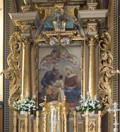 Zdjęcie nr 1: Nastawa ołtarzowa jednoosiowa, jednokondygnacyjna ze zwieńczeniem, ustawiona na wysokim dwustrefowym cokole, zryzalitowanym na zewnetrznych krawędziach. Ołtarz prostopadłościenny z kamienną mensą. Antepedium drewniane, dekorowane na wybrzuszeniu trzema płycinami z rytą wicią roślinną i monogramem IHS. Tabernakulum w kształcie kapliczki na cokole w formie prostopadłościanu; ścianka frontowa ujęta półfilarami dźwigającymi gzyms opasujący go z trzech stron, na froncie przyjmujący kształt łuku segmentowego, spiętego ustawionymi przeciwstawnie wolutami. Trzony półfilarów dekorowane owocowo-roślinnymi zwisami, pole środkowe skręconymi liśćmi akantu w układzie lustrzanym. Na nim nastawa w formie półokrągłej ścianki dekorowanej płyciną w kształcie stojącego prostokąta zamkniętego łukiem pełnym zdobionym kielichem z unoszącą się ponad nim hostią w otoczeniu promieni. Drzwiczki ujęte skręconymi kolumnami dźwigającymi pełne belkowanie wyłamane na ich osiach i wyniesione w łuk na polem centralnym. Na środku fryzu uskrzydlona główka anielska. Na wyłamanych częściach belkowania wazony płomieniste. Nastawa ołtarzowa ujęta parą kolumn o kapitelach kompozytowych dźwigających fragmenty belkowania o silnie wysuniętym gzymsie, połączone architrawem przyjmującym nad polem środkowym kształt łuku pełnego. Pole środkowe niemal w całości wypełnia nisza ujęta ramą w formie stojącego prostokąta wyłamanego prostokątnie w partii górnego boku, wypełniona obrazem św. Rodziny wymiennie z wizerunkiem Matki Boskiej Niepokalanie Poczętej. Zwieńczenie na zwężającej się łukowato podstawie w formie przyczółka przerwanego pośrodku cokołem z krzyżem. Cokół w górnej strefie dekorowany płycinami, w partii ryzalitów wypełnionymi akantem mięsistym i uskrzydloną główką anielską na froncie. Rama obrazu w polu środkowym dekorowana wstęgą i liśćmi; w prostokątnym polu nad obrazem płaskorzeźbione obłoki z rzeźbą Gołębicy Ducha Świętego w glorii; powyżej w polu łuku półpostać Boga Ojca z rozłożonymi na boki rękami. Kondygnacja ujęta uszakami utworzonymi z silnie skręcanego mięsistego akantu, zwiniętego w woluty oraz guzy. Krawędzie podstawy zwieńczenia oraz gzyms przyczółka dekorowane liśćmi akantu. Struktura polichromowana w kolorze kremowym; ornamentyka, profile i kolumny złocone i srebrzone.