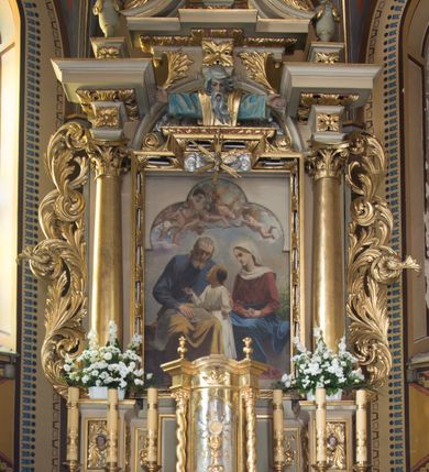 Zdjęcie nr 10: Nastawa ołtarzowa jednoosiowa, jednokondygnacyjna ze zwieńczeniem, ustawiona na wysokim dwustrefowym cokole, zryzalitowanym na zewnetrznych krawędziach. Ołtarz prostopadłościenny z kamienną mensą. Antepedium drewniane, dekorowane na wybrzuszeniu trzema płycinami z rytą wicią roślinną i monogramem IHS. Tabernakulum w kształcie kapliczki na cokole w formie prostopadłościanu; ścianka frontowa ujęta półfilarami dźwigającymi gzyms opasujący go z trzech stron, na froncie przyjmujący kształt łuku segmentowego, spiętego ustawionymi przeciwstawnie wolutami. Trzony półfilarów dekorowane owocowo-roślinnymi zwisami, pole środkowe skręconymi liśćmi akantu w układzie lustrzanym. Na nim nastawa w formie półokrągłej ścianki dekorowanej płyciną w kształcie stojącego prostokąta zamkniętego łukiem pełnym zdobionym kielichem z unoszącą się ponad nim hostią w otoczeniu promieni. Drzwiczki ujęte skręconymi kolumnami dźwigającymi pełne belkowanie wyłamane na ich osiach i wyniesione w łuk na polem centralnym. Na środku fryzu uskrzydlona główka anielska. Na wyłamanych częściach belkowania wazony płomieniste. Nastawa ołtarzowa ujęta parą kolumn o kapitelach kompozytowych dźwigających fragmenty belkowania o silnie wysuniętym gzymsie, połączone architrawem przyjmującym nad polem środkowym kształt łuku pełnego. Pole środkowe niemal w całości wypełnia nisza ujęta ramą w formie stojącego prostokąta wyłamanego prostokątnie w partii górnego boku, wypełniona obrazem św. Rodziny wymiennie z wizerunkiem Matki Boskiej Niepokalanie Poczętej. Zwieńczenie na zwężającej się łukowato podstawie w formie przyczółka przerwanego pośrodku cokołem z krzyżem. Cokół w górnej strefie dekorowany płycinami, w partii ryzalitów wypełnionymi akantem mięsistym i uskrzydloną główką anielską na froncie. Rama obrazu w polu środkowym dekorowana wstęgą i liśćmi; w prostokątnym polu nad obrazem płaskorzeźbione obłoki z rzeźbą Gołębicy Ducha Świętego w glorii; powyżej w polu łuku półpostać Boga Ojca z rozłożonymi na boki rękami. Kondygnacja ujęta uszakami utworzonymi z silnie skręcanego mięsistego akantu, zwiniętego w woluty oraz guzy. Krawędzie podstawy zwieńczenia oraz gzyms przyczółka dekorowane liśćmi akantu. Struktura polichromowana w kolorze kremowym; ornamentyka, profile i kolumny złocone i srebrzone.