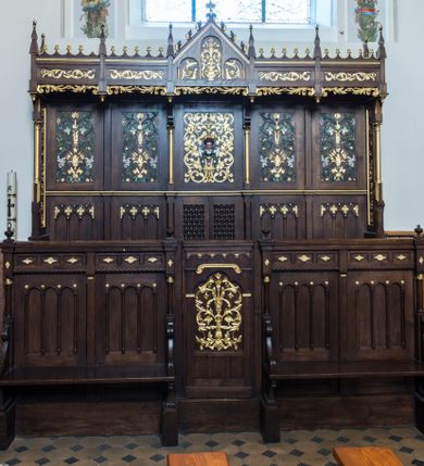 Zdjęcie nr 1: Dębowe, przyścienne stalle uzyskały formę neogotyckiej, pięcioosiowej struktury architektonicznej z zapleckiem i baldachimem. Całość wznosi się na wysokim postumencie. Wejście do nich prowadzi z boków oraz z przodu na osi centralnej, gdzie wprawiono drzwiczki. Ozdobiono je płaskorzeźbioną i złoconą ornamentalną wicią roślinną wpisaną w prostokątną płycinę zwieńczoną stylizowanym trójliściem. Ponad płyciną wprowadzono fryz lombardzki, a poniżej trzy mniejsze, prostokątne, puste płyciny. Na pozostałych osiach na przedpiersiu usytuowano cztery płyciny, które stanowią zaplecek dla dwóch ławek flankujących wejście środkowe. Ławki ujmują wolutowe boki, zdobione stylizowanymi liśćmi akantu, przechodzące poniżej na dekoracyjne wsporniki ustawione na wysokich cokolikach. Jednocześnie wolutowe boki przylegają do wysokich postumentów, na których ustawiono kolumienki o złoconych kapitelach korynckich, podtrzymujących wyłamane belkowanie, stanowiące jednocześnie parapet stalli. Każdą z dolnych płycin zdobi triada półokrągłych arkadek wspartych na smukłych kolumienkach ustawionych na cokole. Także belkowanie artykułują triady płycin ozdobione złoconymi i płaskorzeźbionymi motywami kwiatowymi w romboidalnych obramieniach. Również zaplecek stali uzyskał pięcioosiową artykulację wyznaczoną przez płyciny ułożone w dwóch poziomach. W dolnym poziomie użyto płycin dekorowanych triadami trójkątnych arkadek na smukłych filarkach, które w górnej części tworzą dekoracyjną, romboidalną kratownicę z wpisanym złoconym ornamentem roślinnym. Tą rytmikę przerywa pole osi środkowej, gdzie wprawiono dwuskrzydłowe drzwiczki z ażurową romboidalną kratką. W górnym poziomie wprawiono prostokątne płyciny ozdobione płaskorzeźbioną, polichromowaną i złoconą dekoracją floralno-roślinną, gdzie można wyróżnić złote kłosy zbóż, stylizowane kwiaty lilii górskich i szarotek. Płycinę na osi środkowej  zdobi bujny, płaskorzeźbiony i złocony ornament roślinny, w którego środku widoczna jest uskrzydlona główka aniołka. Jego srebrzone skrzydła przewiązane są zieloną stułą kapłańską. Po bokach płycinę flankują dwie złocone kolumienki o korynckich kapitelach na niskich cokolikach, podtrzymujące wysokie imposty, na które spływają wolutowe konsole. Wspierają one trójkątny przyczółek rozdzielający na dwie części baldachim stalli. Dekoracyjny baldachim wspiera się na wąskich bokach zdobionych od przodu smukłymi, złoconymi kolumienkami o korynckich kapitelach na wysokich postumentach. Za kolumienkami znajdują się płyciny wypełnione ażurowym, złoconym ornamentem roślinnym. Pięcioosiowy podział baldachimu odpowiada artykulacji zaplecka oraz przedpiersi stalli. Tworzy go wypiętrzony trójkątny przyczółek zdobiony czołgankami i kwiatonem oraz cztery prostokątne, wąskie płyciny rozdzielone stylizowanymi pinaklami. Pomiędzy nimi u góry rozciąga się odwrócona arkatura, a u dołu ażurowy, złocony ornament roślinny. Pole przyczółka dekoruje triada półokrągłych płycin. Wpisano w nie stylizowane kwiaty w wazonie. Podłużne płyciny w skrajnych osiach zdobione są złoconą i płaskorzeźbioną wicią roślinną.           