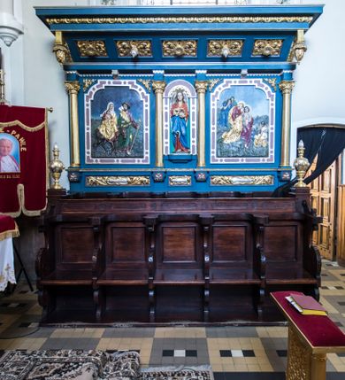 Zdjęcie nr 1: Przyścienne stalle uzyskały formę neobarokowej struktury architektonicznej z zapleckiem i baldachimem. Składają się z lakierowanych i bejcowanych siedzisk na pięcioosowym przedpiersiu oraz pięciu siedzisk pod niebieskim i złoconym baldachimem wspartym na trójosiowym, także niebieskim i złoconym zaplecku. Każde z ruchomych siedzisk przedpiersia po bokach z obu stron zaopatrzone zostało w wycinane z deski wolutowe oparcie, podtrzymujące podkowiaste w wykroju zwieńczenie oparcia. Dodatkowo ruchome siedzisko na rzucie czworoboku wspiera się na wolutowych podporach stanowiących przedłużenie oparć, ustawionych na niskim, drewnianym podwyższeniu. Zaplecki zostały ozdobione czworobocznymi płycinami ujętymi w wąskie, profilowane obramienia. Przedpiersie stalli flankują czworoboczne, smukłe postument na których ustawiono stylizowane wazy płomieniste. W ten sam sposób, co na przedpiersiu, zaaranżowano siedziska pod baldachimem. Za nimi rozciąga się trójosiowy zaplecek, który uzyskał kształt wydłużonego prostokąta, na niewysokim cokole, ozdobionym płycinami w formie leżących prostokątów. Płyciny ozdobiono stylizowanym ornamentem akantowym. Artykulację pionową zaplecka przeprowadzona za pomocą czterech gładkich kolumn z impostami wspierający wyłamujące się na osiach fragmenty belkowania. W polu środkowym znalazła się półokrągła nisza z figurą anioła, trzymającego obiema rękami koronę cierniową. Niszę ujmuje rama utworzona ze stylizowanego ornamentu okuciowego. W polach bocznych zastosowano płyciny w formie stojącego prostokąta o ściętych narożach, w ramach utworzonych ze stylizowanego ornamentu okuciowego. W płycinach umieszczono płaskorzeźby przedstawiające po stronie lewej scenę &quot;Ucieczka do Egiptu&quot;, po stronie prawej &quot;Złożenie do grobu&quot;. W podniebiu baladachimu znalazło się pięć kasetonowych płycin, naprzemiennie zdobionych kwiatowymi rozetkami i srebrzonymi szyszkami. W partii fryzu dekorującego baldachim użyto ornamentu utworzonego z wolich oczu. Zewnętrzne boki baldachimu wsparto na wolucie zakończonej od przodu wiszącymi złoconymi szyszkami. Wieńczący go gzyms jest profilowany i złocon. Anioł w niszy ukazany został w pozie lekkiego kontrapostu, w dłoniach trzyma na wysokości piersi złoconą koronę cierniową. Jego twarz okalają długie, brązowe włosy spływające na ramiona. Odziany jest w długą czerwoną tunikę, z wywijającą się jasnobeżową podszewką. Przy szyi zdobi ją złota lamówka. Wokół bioder ma przewiązany niebieski płaszcz o jasnej podszewce. Z tyłu postaci widoczne są srebrne, długie, złożone skrzydła. Płaskorzeźba po stronie lewej w zaplecku przedstawia ucieczkę Świętej Rodziny do Egiptu. W centralnym punkcie kompozycji ukazano jadącą na osiołku Matkę Boską, trzymającą na rękach Dzieciątko. Obok idzie św. Józef. Madonna siedzi na osiołku frontalnie z głową ujętą w trzech czwartych w prawo. Ma podłużną twarz o jasnej karnacji z półprzymkniętymi powiekami, prosty nos i wydatne usta. Ubrana jest w czerwoną suknię i złoty płaszcz, narzucony na lewe ramie, spływający na kolana i okrywający nóżki nagiego Dzieciątka trzymanego na kolanach. Na głowie Maria ma białą chustę, której lewy koniec osłania szyję i opada na prawe ramię. Chusta na czole odsłania fragment brązowych włosów. Siedzący Jezus został ukazany w trzech czwartych w prawo. Ma owalną twarz o jasnej karnacji i rumianych policzkach, drobny nos i wargi. Głowę otaczają krótkie, brązowe włosy. Dolną część torsu Dzieciątka owija biała chusta, natomiast nogi od kolan przykrywa złoty płaszcz Marii. Idący przodem św. Józef ukazany został w całej postaci z głową odwróconą w stronę Matki Boskiej. W wysuniętej do przodu lewej ręce trzyma zakrzywioną laskę, a w prawej zgiętej w łokciu lejce. Święty ma pociągłą twarz okoloną brodą oraz włosami częściowo widocznymi spod kapelusza o podwiniętym rondzie. Ubrany jest w bordową tunikę odsłaniającą stopy obute w sandały i przerzucony przez prawe ramię zielony płaszcz z wywiniętą, złotą podszewką. Z tyłu na plecach niesie siekierę z ostrzem zwróconym w prawo. Osioł przedstawiony został w profilu o ciemnobrązowej maści. Postacie zostały ukazane na tle ubogiego krajobrazu oraz niebieskiego nieba. Płaskorzeźba po stronie prawej przedstawiając złożenie do grobu, na pierwszym planie wyobraża podtrzymywaną  przez św. Jana Matkę Boską. Stojąca na niewielkim wzniesieniu Madonna ujęta została w trzech czwartych w prawo. Lewą ręką opiera się na ramieniu św. Jana. Prawą rękę ma opuszczoną bezwładnie wzdłuż tułowia. Głowę przechyla w lewo, kierując wzrok w dół. Ma jasną karnację, prosty nos i półprzymknięte powieki. Ubrana jest w złoty maforion, którego mocno drapowane poły ma przewiązane w pasie. Spod niego widać obszerną, bordową suknię przewiązaną w talii. Schodzący ze wzniesienia św. Jan prawym ramieniem obejmuje Marię, lewą zaś przyciśniętą do piersi trzyma fragment płaszcza. Głowę nieznacznie ma przechyloną w kierunku Marii. Jego młodzieńczą twarz o jasnej karnacji okalają krótkie, brązowe włosy. Apostoł ubrany jest w ciemnobeżową luźną tunikę przewiązaną w pasie i narzucony na lewe ramię bordowy płaszcz o wywijającej się srebrnej podszewce. Wokół głów obu postaci widoczne są nimby. W tle po lewej stronie widoczne są dwie płaczące Marie. Jedna ukazana z dłońmi zakrywającymi twarz, odziana jest w niebieską szatę, przewiązaną w pasie. Ma długie, rozpuszczone brązowe włosy. Druga kobieta ubrana jest w zielony maforion założony na piersi. Za nimi w tle widoczny jest szczyt Golgoty z dwoma krzyżami. Po stronie prawej, w dolnym rogu przedstawienia, ukazano dwóch mężczyzn niosących ciało zmarłego Chrystusa, owinięte w złoty całun. Widać tylko z profilu twarz Jezusa o brązowym zaroście i włosach spływających na ramiona. Jeden z niosących ciało mężczyzn ukazany jest od tyłu, w trzech czwartych w lewo. Obiema rękami trzyma zmarłego pod ramionami. Jego głowa otoczona krótkimi włosami i brązowym zarostem jest widoczna z profilu. Mężczyzna ubrany jest w krótką, srebrną tunikę sięgającą kolan i przewiązaną w pasie. Druga postać  odwrócona do widza tyłem trzyma z prawej strony obie nogi Zbawiciela. Jest to mężczyzna o krótkich, brązowych włosach w niebieskiej szacie. Przed nimi w tle widać zarys miejskiej architektury i skaliste gór.
