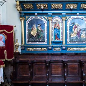 Zdjęcie nr 1: Przyścienne stalle uzyskały formę neobarokowej struktury architektonicznej z zapleckiem i baldachimem. Składają się z lakierowanych i bejcowanych siedzisk na pięcioosowym przedpiersiu oraz pięciu siedzisk pod niebieskim i złoconym baldachimem wspartym na trójosiowym, także niebieskim i złoconym zaplecku. Każde z ruchomych siedzisk przedpiersia po bokach z obu stron zaopatrzone zostało w wycinane z deski wolutowe oparcie, podtrzymujące podkowiaste w wykroju zwieńczenie oparcia. Dodatkowo ruchome siedzisko na rzucie czworoboku wspiera się na wolutowych podporach stanowiących przedłużenie oparć, ustawionych na niskim, drewnianym podwyższeniu. Zaplecki zostały ozdobione czworobocznymi płycinami ujętymi w wąskie, profilowane obramienia. Przedpiersie stalli flankują czworoboczne, smukłe postument na których ustawiono stylizowane wazy płomieniste. W ten sam sposób, co na przedpiersiu, zaaranżowano siedziska pod baldachimem. Za nimi rozciąga się trójosiowy zaplecek, który uzyskał kształt wydłużonego prostokąta, na niewysokim cokole, ozdobionym płycinami w formie leżących prostokątów. Płyciny ozdobiono stylizowanym ornamentem akantowym. Artykulację pionową zaplecka przeprowadzona za pomocą czterech gładkich kolumn z impostami wspierający wyłamujące się na osiach fragmenty belkowania. W polu środkowym znalazła się półokrągła nisza z figurą anioła, trzymającego obiema rękami koronę cierniową. Niszę ujmuje rama utworzona ze stylizowanego ornamentu okuciowego. W polach bocznych zastosowano płyciny w formie stojącego prostokąta o ściętych narożach, w ramach utworzonych ze stylizowanego ornamentu okuciowego. W płycinach umieszczono płaskorzeźby przedstawiające po stronie lewej scenę "Ucieczka do Egiptu", po stronie prawej "Złożenie do grobu". W podniebiu baladachimu znalazło się pięć kasetonowych płycin, naprzemiennie zdobionych kwiatowymi rozetkami i srebrzonymi szyszkami. W partii fryzu dekorującego baldachim użyto ornamentu utworzonego z wolich oczu. Zewnętrzne boki baldachimu wsparto na wolucie zakończonej od przodu wiszącymi złoconymi szyszkami. Wieńczący go gzyms jest profilowany i złocon. Anioł w niszy ukazany został w pozie lekkiego kontrapostu, w dłoniach trzyma na wysokości piersi złoconą koronę cierniową. Jego twarz okalają długie, brązowe włosy spływające na ramiona. Odziany jest w długą czerwoną tunikę, z wywijającą się jasnobeżową podszewką. Przy szyi zdobi ją złota lamówka. Wokół bioder ma przewiązany niebieski płaszcz o jasnej podszewce. Z tyłu postaci widoczne są srebrne, długie, złożone skrzydła. Płaskorzeźba po stronie lewej w zaplecku przedstawia ucieczkę Świętej Rodziny do Egiptu. W centralnym punkcie kompozycji ukazano jadącą na osiołku Matkę Boską, trzymającą na rękach Dzieciątko. Obok idzie św. Józef. Madonna siedzi na osiołku frontalnie z głową ujętą w trzech czwartych w prawo. Ma podłużną twarz o jasnej karnacji z półprzymkniętymi powiekami, prosty nos i wydatne usta. Ubrana jest w czerwoną suknię i złoty płaszcz, narzucony na lewe ramie, spływający na kolana i okrywający nóżki nagiego Dzieciątka trzymanego na kolanach. Na głowie Maria ma białą chustę, której lewy koniec osłania szyję i opada na prawe ramię. Chusta na czole odsłania fragment brązowych włosów. Siedzący Jezus został ukazany w trzech czwartych w prawo. Ma owalną twarz o jasnej karnacji i rumianych policzkach, drobny nos i wargi. Głowę otaczają krótkie, brązowe włosy. Dolną część torsu Dzieciątka owija biała chusta, natomiast nogi od kolan przykrywa złoty płaszcz Marii. Idący przodem św. Józef ukazany został w całej postaci z głową odwróconą w stronę Matki Boskiej. W wysuniętej do przodu lewej ręce trzyma zakrzywioną laskę, a w prawej zgiętej w łokciu lejce. Święty ma pociągłą twarz okoloną brodą oraz włosami częściowo widocznymi spod kapelusza o podwiniętym rondzie. Ubrany jest w bordową tunikę odsłaniającą stopy obute w sandały i przerzucony przez prawe ramię zielony płaszcz z wywiniętą, złotą podszewką. Z tyłu na plecach niesie siekierę z ostrzem zwróconym w prawo. Osioł przedstawiony został w profilu o ciemnobrązowej maści. Postacie zostały ukazane na tle ubogiego krajobrazu oraz niebieskiego nieba. Płaskorzeźba po stronie prawej przedstawiając złożenie do grobu, na pierwszym planie wyobraża podtrzymywaną  przez św. Jana Matkę Boską. Stojąca na niewielkim wzniesieniu Madonna ujęta została w trzech czwartych w prawo. Lewą ręką opiera się na ramieniu św. Jana. Prawą rękę ma opuszczoną bezwładnie wzdłuż tułowia. Głowę przechyla w lewo, kierując wzrok w dół. Ma jasną karnację, prosty nos i półprzymknięte powieki. Ubrana jest w złoty maforion, którego mocno drapowane poły ma przewiązane w pasie. Spod niego widać obszerną, bordową suknię przewiązaną w talii. Schodzący ze wzniesienia św. Jan prawym ramieniem obejmuje Marię, lewą zaś przyciśniętą do piersi trzyma fragment płaszcza. Głowę nieznacznie ma przechyloną w kierunku Marii. Jego młodzieńczą twarz o jasnej karnacji okalają krótkie, brązowe włosy. Apostoł ubrany jest w ciemnobeżową luźną tunikę przewiązaną w pasie i narzucony na lewe ramię bordowy płaszcz o wywijającej się srebrnej podszewce. Wokół głów obu postaci widoczne są nimby. W tle po lewej stronie widoczne są dwie płaczące Marie. Jedna ukazana z dłońmi zakrywającymi twarz, odziana jest w niebieską szatę, przewiązaną w pasie. Ma długie, rozpuszczone brązowe włosy. Druga kobieta ubrana jest w zielony maforion założony na piersi. Za nimi w tle widoczny jest szczyt Golgoty z dwoma krzyżami. Po stronie prawej, w dolnym rogu przedstawienia, ukazano dwóch mężczyzn niosących ciało zmarłego Chrystusa, owinięte w złoty całun. Widać tylko z profilu twarz Jezusa o brązowym zaroście i włosach spływających na ramiona. Jeden z niosących ciało mężczyzn ukazany jest od tyłu, w trzech czwartych w lewo. Obiema rękami trzyma zmarłego pod ramionami. Jego głowa otoczona krótkimi włosami i brązowym zarostem jest widoczna z profilu. Mężczyzna ubrany jest w krótką, srebrną tunikę sięgającą kolan i przewiązaną w pasie. Druga postać  odwrócona do widza tyłem trzyma z prawej strony obie nogi Zbawiciela. Jest to mężczyzna o krótkich, brązowych włosach w niebieskiej szacie. Przed nimi w tle widać zarys miejskiej architektury i skaliste gór.