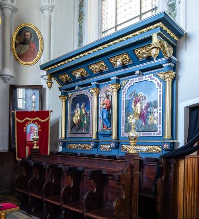 Zdjęcie nr 2: Przyścienne stalle uzyskały formę neobarokowej struktury architektonicznej z zapleckiem i baldachimem. Składają się z lakierowanych i bejcowanych siedzisk na pięcioosowym przedpiersiu oraz pięciu siedzisk pod niebieskim i złoconym baldachimem wspartym na trójosiowym, także niebieskim i złoconym zaplecku. Każde z ruchomych siedzisk przedpiersia po bokach z obu stron zaopatrzone zostało w wycinane z deski wolutowe oparcie, podtrzymujące podkowiaste w wykroju zwieńczenie oparcia. Dodatkowo ruchome siedzisko na rzucie czworoboku wspiera się na wolutowych podporach stanowiących przedłużenie oparć, ustawionych na niskim, drewnianym podwyższeniu. Zaplecki zostały ozdobione czworobocznymi płycinami ujętymi w wąskie, profilowane obramienia. Przedpiersie stalli flankują czworoboczne, smukłe postument na których ustawiono stylizowane wazy płomieniste. W ten sam sposób, co na przedpiersiu, zaaranżowano siedziska pod baldachimem. Za nimi rozciąga się trójosiowy zaplecek, który uzyskał kształt wydłużonego prostokąta, na niewysokim cokole, ozdobionym płycinami w formie leżących prostokątów. Płyciny ozdobiono stylizowanym ornamentem akantowym. Artykulację pionową zaplecka przeprowadzona za pomocą czterech gładkich kolumn z impostami wspierający wyłamujące się na osiach fragmenty belkowania. W polu środkowym znalazła się półokrągła nisza z figurą anioła, trzymającego obiema rękami koronę cierniową. Niszę ujmuje rama utworzona ze stylizowanego ornamentu okuciowego. W polach bocznych zastosowano płyciny w formie stojącego prostokąta o ściętych narożach, w ramach utworzonych ze stylizowanego ornamentu okuciowego. W płycinach umieszczono płaskorzeźby przedstawiające po stronie lewej scenę "Ucieczka do Egiptu", po stronie prawej "Złożenie do grobu". W podniebiu baladachimu znalazło się pięć kasetonowych płycin, naprzemiennie zdobionych kwiatowymi rozetkami i srebrzonymi szyszkami. W partii fryzu dekorującego baldachim użyto ornamentu utworzonego z wolich oczu. Zewnętrzne boki baldachimu wsparto na wolucie zakończonej od przodu wiszącymi złoconymi szyszkami. Wieńczący go gzyms jest profilowany i złocon. Anioł w niszy ukazany został w pozie lekkiego kontrapostu, w dłoniach trzyma na wysokości piersi złoconą koronę cierniową. Jego twarz okalają długie, brązowe włosy spływające na ramiona. Odziany jest w długą czerwoną tunikę, z wywijającą się jasnobeżową podszewką. Przy szyi zdobi ją złota lamówka. Wokół bioder ma przewiązany niebieski płaszcz o jasnej podszewce. Z tyłu postaci widoczne są srebrne, długie, złożone skrzydła. Płaskorzeźba po stronie lewej w zaplecku przedstawia ucieczkę Świętej Rodziny do Egiptu. W centralnym punkcie kompozycji ukazano jadącą na osiołku Matkę Boską, trzymającą na rękach Dzieciątko. Obok idzie św. Józef. Madonna siedzi na osiołku frontalnie z głową ujętą w trzech czwartych w prawo. Ma podłużną twarz o jasnej karnacji z półprzymkniętymi powiekami, prosty nos i wydatne usta. Ubrana jest w czerwoną suknię i złoty płaszcz, narzucony na lewe ramie, spływający na kolana i okrywający nóżki nagiego Dzieciątka trzymanego na kolanach. Na głowie Maria ma białą chustę, której lewy koniec osłania szyję i opada na prawe ramię. Chusta na czole odsłania fragment brązowych włosów. Siedzący Jezus został ukazany w trzech czwartych w prawo. Ma owalną twarz o jasnej karnacji i rumianych policzkach, drobny nos i wargi. Głowę otaczają krótkie, brązowe włosy. Dolną część torsu Dzieciątka owija biała chusta, natomiast nogi od kolan przykrywa złoty płaszcz Marii. Idący przodem św. Józef ukazany został w całej postaci z głową odwróconą w stronę Matki Boskiej. W wysuniętej do przodu lewej ręce trzyma zakrzywioną laskę, a w prawej zgiętej w łokciu lejce. Święty ma pociągłą twarz okoloną brodą oraz włosami częściowo widocznymi spod kapelusza o podwiniętym rondzie. Ubrany jest w bordową tunikę odsłaniającą stopy obute w sandały i przerzucony przez prawe ramię zielony płaszcz z wywiniętą, złotą podszewką. Z tyłu na plecach niesie siekierę z ostrzem zwróconym w prawo. Osioł przedstawiony został w profilu o ciemnobrązowej maści. Postacie zostały ukazane na tle ubogiego krajobrazu oraz niebieskiego nieba. Płaskorzeźba po stronie prawej przedstawiając złożenie do grobu, na pierwszym planie wyobraża podtrzymywaną  przez św. Jana Matkę Boską. Stojąca na niewielkim wzniesieniu Madonna ujęta została w trzech czwartych w prawo. Lewą ręką opiera się na ramieniu św. Jana. Prawą rękę ma opuszczoną bezwładnie wzdłuż tułowia. Głowę przechyla w lewo, kierując wzrok w dół. Ma jasną karnację, prosty nos i półprzymknięte powieki. Ubrana jest w złoty maforion, którego mocno drapowane poły ma przewiązane w pasie. Spod niego widać obszerną, bordową suknię przewiązaną w talii. Schodzący ze wzniesienia św. Jan prawym ramieniem obejmuje Marię, lewą zaś przyciśniętą do piersi trzyma fragment płaszcza. Głowę nieznacznie ma przechyloną w kierunku Marii. Jego młodzieńczą twarz o jasnej karnacji okalają krótkie, brązowe włosy. Apostoł ubrany jest w ciemnobeżową luźną tunikę przewiązaną w pasie i narzucony na lewe ramię bordowy płaszcz o wywijającej się srebrnej podszewce. Wokół głów obu postaci widoczne są nimby. W tle po lewej stronie widoczne są dwie płaczące Marie. Jedna ukazana z dłońmi zakrywającymi twarz, odziana jest w niebieską szatę, przewiązaną w pasie. Ma długie, rozpuszczone brązowe włosy. Druga kobieta ubrana jest w zielony maforion założony na piersi. Za nimi w tle widoczny jest szczyt Golgoty z dwoma krzyżami. Po stronie prawej, w dolnym rogu przedstawienia, ukazano dwóch mężczyzn niosących ciało zmarłego Chrystusa, owinięte w złoty całun. Widać tylko z profilu twarz Jezusa o brązowym zaroście i włosach spływających na ramiona. Jeden z niosących ciało mężczyzn ukazany jest od tyłu, w trzech czwartych w lewo. Obiema rękami trzyma zmarłego pod ramionami. Jego głowa otoczona krótkimi włosami i brązowym zarostem jest widoczna z profilu. Mężczyzna ubrany jest w krótką, srebrną tunikę sięgającą kolan i przewiązaną w pasie. Druga postać  odwrócona do widza tyłem trzyma z prawej strony obie nogi Zbawiciela. Jest to mężczyzna o krótkich, brązowych włosach w niebieskiej szacie. Przed nimi w tle widać zarys miejskiej architektury i skaliste gór.