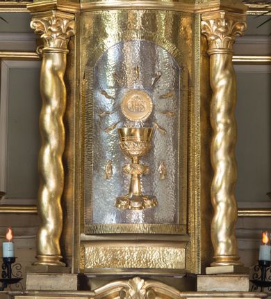 Zdjęcie nr 1: Tabernakulum oparte na schemacie aediculi  na cokole w formie prostopadłościanu. Ścianka frontowa ujęta półfilarami dźwigającymi gzyms opasujący go z trzech stron, na froncie przyjmujący kształt łuku segmentowego, spiętego ustawionymi przeciwstawnie wolutami. Trzony półfilarów dekorowane owocowo-roślinnymi zwisami, pole środkowe skręconymi liśćmi akantu w układzie lustrzanym. Na nim nastawa w formie półokrągłej ścianki dekorowanej płyciną w kształcie stojącego prostokąta zamkniętego łukiem pełnym zdobionym kielichem z unoszącą się ponad nim hostią w otoczeniu promieni. Drzwiczki ujęte skręconymi kolumnami dźwigającymi pełne belkowanie wyłamane na ich osiach i wyniesione w łuk na polem centralnym. Na środku fryzu uskrzydlona główka anielska. Na wyłamanych częściach belkowania wazony płomieniste. Na ściance bocznej tabliczka z inskrypcją „FUNDATORZY / RODZICE I DZIECI I szej KOMUNI ŚW(IĘTEJ) / ROCZNIK 2002 / WYK(ONANE) KRAKÓW / MAJ 2002 R(OKU)”. 