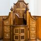 Zdjęcie nr 1: Przyścienny, otwarty konfesjonał szafiasty, wykonany w całości z drewna uzyskał formę architektoniczną ustawioną na niskim cokole. Drewniane siedzisko z podłokietnikami i wysokim zapleckiem flankują dwie ścianki boczne przeprute dwoma kwadratowymi okienkami wypełnionymi romboidalną kratownicą. Ścianki od góry zamknięte są wyciętym, odwróconym łukiem odcinkowym. Od frontu zamykają je smukłe czworoboczne filarki zwieńczone zdwojonymi płaskimi kulami z krzyżykami. W dolnej partii do filarków od przodu przymocowano półwypukłe drzwi. Zdobią je cztery kwadratowe płyciny, obramione profilowanymi listwami. Przestrzeń pomiędzy płycinami tworzy krzyż równoramienny. W górnej części do filarków przymocowano dwuskrzydłowe drzwiczki, których przody ozdobiono płaskorzeźbionym motywem stylizowanego dziewięciosiłu. Podobny motyw wpisano w pola ułożonych w pionowym rzędzie trzech płycin obramionych profilowanymi listwami. Płyciny umieszczono na bocznych policzkach konfesjonału. Górne płyciny uzyskały kształt stojącego trapezu, natomiast pozostałe cztery mają kształt czworoboczny. Policzki wieńczą wolutowe spływy. Zaplecek konfesjonału w górnej części ozdobiony został czworoboczną płyciną o wklęsłych narożach, ujętą szeroką, wklęsło-wypukłą ramą. Pole płyciny ozdobiono krzyżem utworzonym z fazowanych listew, w kształcie zbliżonym do krzyża równoramiennego z wpisanym okręgiem.   