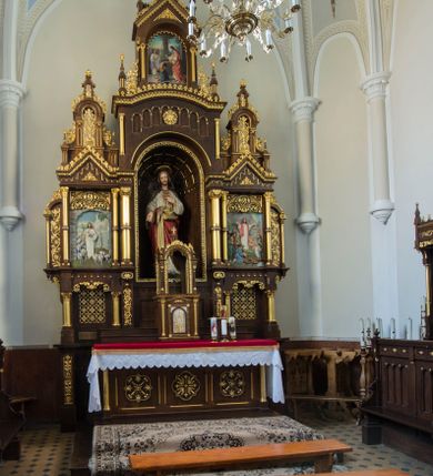Zdjęcie nr 2: Przyścienny trójosiowy, jednokondygnacyjny ołtarz architektoniczny ustawiono na dwustopniowym podeście. Stipes ołtarzowy flankowany przez dwie złocone kolumienki wspierające mensę, ozdobiono trzema płycinami zamkniętymi łukiem odcinkowym. Pola skrajne płycin wypełnia romboidalny czworobok wpisany w owal. Jego wnętrze dekoruje ornamentyka geometryczno-roślinna. Pole środkowe ozdobiono kwadratem wpisanym w owal, w środku którego umieszczono krzyż równoramienny w promienistym otoku. Na mensie ustawiono czworoboczne tabernakulum zwieńczone baldachimem. Front tabernakulum flankują dwie, smukłe, złocone kolumienki o korynckich kapitelach podtrzymujące spłaszczony łuk odcinkowy. Srebrzone półokrągłe drzwiczki wpisano w prostokątne obramienie dekorowane w narożach złoconymi rozetkami. Na drzwiczkach umieszczono chrystogram "IHS" ponad kłosami i winnym gronem. Baldachim tabernakulum uzyskał formę ażurowej, czworobocznej kapliczki o półkoliście zamkniętych, prostokątnych ściankach z wpisanym w górnej części trójliściem. W jej narożach umieszczono kolumienki na wysokich postumentach z kapitelami kostkowymi. Trójosiowe retabulum wsparto na wysokim postumencie, flankowanym przez nieznacznie wysunięte smukłe podpory, dekorowane od frontu płaskorzeźbionym ornamentem roślinnym. Na nich wspierają się skrajne kolumienki, zamykające partię cokołową nastawy. Artykułują je dwie edikule w skrajnych osiach zamknięte łukiem odcinkowym, ozdobionym płaskorzeźbionym ornamentem roślinnym. Wnętrze edikul wypełnia złocona kratownica z polami zdobionymi motywem stylizowanego krzyżyka. Ponad nimi widoczny jest fryz arkadkowy. Na osi środkowej umieszczono prostokątną, półkoliście zamknietą wnękę o konsze dekorowanej stylizowanymi kasetonami. W niej ustawiono stojacą figurę Najświętszego Serca Pana Jezusa. Wnękę flankują dwie złocone kolumny o kapitelach kostkowych i trzonach zdobionych pierścieniami. Podtrzymują one nadbudowę ze zwieńczeniem zakończonym latarnią. Nadbudowę u góry zamknięto nadwieszonym łukiem odcinkowym. Po bokach flankują ją dwie złocone kolumienki o korynckich kapitelach. Pomiędzy kolumnami widoczny jest fryz arkadowy z umieszczoną na środku złoconą rozetą. Łuk w górnej partii udekorowano złoconą koronką oraz znajdującymi się po bokach dwiema, wielobocznymi sterczynami. Zwieńczenie uzyskało kształt płaskiej kapliczki o dwuspadowym dachu z prostokątnym, półkoliście zamkniętym polem środkowym flankowanym parą kolumienek. Pole dekorowane arkaturą wypełnia obraz "Powrót syna Marnotrawnego". Na dachu kapliczki ustawiono na osi ażurową latarnię a po jej bokach wieloboczne sterczyny. Ponad skrajnymi edikulami w osiach bocznych w partii cokołowej usytuowano płyciny zamknięte po bokach złoconymi kolumienkami z kapitelami kostkowymi, o trzonach dekorowanych pierścieniami, podtrzymujące trójkątne, nadwieszone przyczółki. Zdobi je złocona koronka oraz motyw fryzu arkadkowego. Nad przyczółkami ustawiono zwieńczenie w formie płaskiej kapliczki o półkoliście zamkniętym, ażurowym wykroju wypełnionym złoconym, ornamentem roślinnym z latarenką. Prostokątne płyciny dekorowane w górnej części stylizowanym maswerkiem zawierają płaskorzeźbione przedstawienia. Z lewej strony wyobrażono Chrystusa jako Dobrego Pasterza, a z prawej cudowne rozmnożenie chleba.             