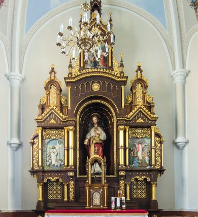 Zdjęcie nr 1: Przyścienny trójosiowy, jednokondygnacyjny ołtarz architektoniczny ustawiono na dwustopniowym podeście. Stipes ołtarzowy flankowany przez dwie złocone kolumienki wspierające mensę, ozdobiono trzema płycinami zamkniętymi łukiem odcinkowym. Pola skrajne płycin wypełnia romboidalny czworobok wpisany w owal. Jego wnętrze dekoruje ornamentyka geometryczno-roślinna. Pole środkowe ozdobiono kwadratem wpisanym w owal, w środku którego umieszczono krzyż równoramienny w promienistym otoku. Na mensie ustawiono czworoboczne tabernakulum zwieńczone baldachimem. Front tabernakulum flankują dwie, smukłe, złocone kolumienki o korynckich kapitelach podtrzymujące spłaszczony łuk odcinkowy. Srebrzone półokrągłe drzwiczki wpisano w prostokątne obramienie dekorowane w narożach złoconymi rozetkami. Na drzwiczkach umieszczono chrystogram "IHS" ponad kłosami i winnym gronem. Baldachim tabernakulum uzyskał formę ażurowej, czworobocznej kapliczki o półkoliście zamkniętych, prostokątnych ściankach z wpisanym w górnej części trójliściem. W jej narożach umieszczono kolumienki na wysokich postumentach z kapitelami kostkowymi. Trójosiowe retabulum wsparto na wysokim postumencie, flankowanym przez nieznacznie wysunięte smukłe podpory, dekorowane od frontu płaskorzeźbionym ornamentem roślinnym. Na nich wspierają się skrajne kolumienki, zamykające partię cokołową nastawy. Artykułują je dwie edikule w skrajnych osiach zamknięte łukiem odcinkowym, ozdobionym płaskorzeźbionym ornamentem roślinnym. Wnętrze edikul wypełnia złocona kratownica z polami zdobionymi motywem stylizowanego krzyżyka. Ponad nimi widoczny jest fryz arkadkowy. Na osi środkowej umieszczono prostokątną, półkoliście zamknietą wnękę o konsze dekorowanej stylizowanymi kasetonami. W niej ustawiono stojacą figurę Najświętszego Serca Pana Jezusa. Wnękę flankują dwie złocone kolumny o kapitelach kostkowych i trzonach zdobionych pierścieniami. Podtrzymują one nadbudowę ze zwieńczeniem zakończonym latarnią. Nadbudowę u góry zamknięto nadwieszonym łukiem odcinkowym. Po bokach flankują ją dwie złocone kolumienki o korynckich kapitelach. Pomiędzy kolumnami widoczny jest fryz arkadowy z umieszczoną na środku złoconą rozetą. Łuk w górnej partii udekorowano złoconą koronką oraz znajdującymi się po bokach dwiema, wielobocznymi sterczynami. Zwieńczenie uzyskało kształt płaskiej kapliczki o dwuspadowym dachu z prostokątnym, półkoliście zamkniętym polem środkowym flankowanym parą kolumienek. Pole dekorowane arkaturą wypełnia obraz "Powrót syna Marnotrawnego". Na dachu kapliczki ustawiono na osi ażurową latarnię a po jej bokach wieloboczne sterczyny. Ponad skrajnymi edikulami w osiach bocznych w partii cokołowej usytuowano płyciny zamknięte po bokach złoconymi kolumienkami z kapitelami kostkowymi, o trzonach dekorowanych pierścieniami, podtrzymujące trójkątne, nadwieszone przyczółki. Zdobi je złocona koronka oraz motyw fryzu arkadkowego. Nad przyczółkami ustawiono zwieńczenie w formie płaskiej kapliczki o półkoliście zamkniętym, ażurowym wykroju wypełnionym złoconym, ornamentem roślinnym z latarenką. Prostokątne płyciny dekorowane w górnej części stylizowanym maswerkiem zawierają płaskorzeźbione przedstawienia. Z lewej strony wyobrażono Chrystusa jako Dobrego Pasterza, a z prawej cudowne rozmnożenie chleba.             