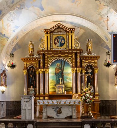 Zdjęcie nr 1: Przyścienną nastawę architektoniczną, trójosiową wykonano z drewna modrzewiowego. Jest to konstrukcja jednokondygnacyjna ze zwieńczeniem, w formie łuku triumfalnego, ustawiona na dwustrefowym cokole. Oś środkową wydzielają dwie pary kolumn o kapitelach kompozytowych i kanelowanych trzonach dźwigających wydłużone imposty. Kolumny wewnętrzne zostały połączone nad polem środkowym fryzem i gzymsem. Z kolei kolumny zewnętrze podtrzymują belkowanie zwieńczenia. Pole środkowe wypełnia nisza w formie stojącego prostokąta zamkniętego łukiem półkolistym z arkaturą z obrazem św. Andrzeja Apostoła. W osiach zewnętrznych usytuowano niższe flankowane parą kanelowanych kolumn z kapitelami kompozytowymi podtrzymujących imposty połączone nad polem fryzem i gzymsem. Nisze uzyskały kształt stojącego prostokąta zamkniętego łukiem półkolistym z arkaturą z figurami św. Wincentego a Paulo i św. Jana Kantego. Ponad niszami na belkowaniu usytuowano cokoły z figurami aniołów trzymających insygnia królewskie. Zwieńczenie uzyskało kształt leżącego prostokąta flankowanego dwiema parami złoconych kolumienek, dźwigających imposty i gzyms nad polem środkowym wyłamanym w trójkąt. Po bokach widoczne są spływy wolutowe. W polu znajduje się rama w formie kwadratu z wpisanym kołem z płaskorzeźbionym przedstawieniem Trójcy Świętej. Cokoły i imposty dekorowane zostały stylizowaną ornamentyką roślinną w postaci zwisów, kampanuli i girland. Struktura nastawy utrzymana jest w kolorze naturalnego drewna, natomiast ornamentyka, detale i profile zostały wyzłocone i wysrebrzone. Z kolei figury i płaskorzeźba w zwieńczeniu uzyskały naturalistyczną polichromię.