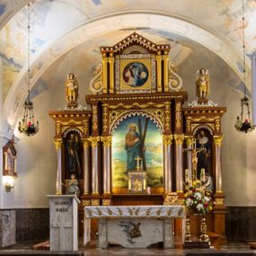 Zdjęcie nr 1: Przyścienną nastawę architektoniczną, trójosiową wykonano z drewna modrzewiowego. Jest to konstrukcja jednokondygnacyjna ze zwieńczeniem, w formie łuku triumfalnego, ustawiona na dwustrefowym cokole. Oś środkową wydzielają dwie pary kolumn o kapitelach kompozytowych i kanelowanych trzonach dźwigających wydłużone imposty. Kolumny wewnętrzne zostały połączone nad polem środkowym fryzem i gzymsem. Z kolei kolumny zewnętrze podtrzymują belkowanie zwieńczenia. Pole środkowe wypełnia nisza w formie stojącego prostokąta zamkniętego łukiem półkolistym z arkaturą z obrazem św. Andrzeja Apostoła. W osiach zewnętrznych usytuowano niższe flankowane parą kanelowanych kolumn z kapitelami kompozytowymi podtrzymujących imposty połączone nad polem fryzem i gzymsem. Nisze uzyskały kształt stojącego prostokąta zamkniętego łukiem półkolistym z arkaturą z figurami św. Wincentego a Paulo i św. Jana Kantego. Ponad niszami na belkowaniu usytuowano cokoły z figurami aniołów trzymających insygnia królewskie. Zwieńczenie uzyskało kształt leżącego prostokąta flankowanego dwiema parami złoconych kolumienek, dźwigających imposty i gzyms nad polem środkowym wyłamanym w trójkąt. Po bokach widoczne są spływy wolutowe. W polu znajduje się rama w formie kwadratu z wpisanym kołem z płaskorzeźbionym przedstawieniem Trójcy Świętej. Cokoły i imposty dekorowane zostały stylizowaną ornamentyką roślinną w postaci zwisów, kampanuli i girland. Struktura nastawy utrzymana jest w kolorze naturalnego drewna, natomiast ornamentyka, detale i profile zostały wyzłocone i wysrebrzone. Z kolei figury i płaskorzeźba w zwieńczeniu uzyskały naturalistyczną polichromię.