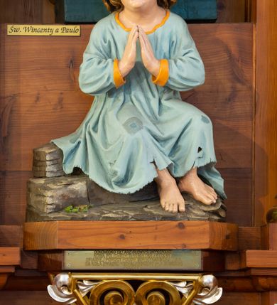 Zdjęcie nr 13: Przyścienną nastawę architektoniczną, trójosiową wykonano z drewna modrzewiowego. Jest to konstrukcja jednokondygnacyjna ze zwieńczeniem, w formie łuku triumfalnego, ustawiona na dwustrefowym cokole. Oś środkową wydzielają dwie pary kolumn o kapitelach kompozytowych i kanelowanych trzonach dźwigających wydłużone imposty. Kolumny wewnętrzne zostały połączone nad polem środkowym fryzem i gzymsem. Z kolei kolumny zewnętrze podtrzymują belkowanie zwieńczenia. Pole środkowe wypełnia nisza w formie stojącego prostokąta zamkniętego łukiem półkolistym z arkaturą z obrazem św. Andrzeja Apostoła. W osiach zewnętrznych usytuowano niższe flankowane parą kanelowanych kolumn z kapitelami kompozytowymi podtrzymujących imposty połączone nad polem fryzem i gzymsem. Nisze uzyskały kształt stojącego prostokąta zamkniętego łukiem półkolistym z arkaturą z figurami św. Wincentego a Paulo i św. Jana Kantego. Ponad niszami na belkowaniu usytuowano cokoły z figurami aniołów trzymających insygnia królewskie. Zwieńczenie uzyskało kształt leżącego prostokąta flankowanego dwiema parami złoconych kolumienek, dźwigających imposty i gzyms nad polem środkowym wyłamanym w trójkąt. Po bokach widoczne są spływy wolutowe. W polu znajduje się rama w formie kwadratu z wpisanym kołem z płaskorzeźbionym przedstawieniem Trójcy Świętej. Cokoły i imposty dekorowane zostały stylizowaną ornamentyką roślinną w postaci zwisów, kampanuli i girland. Struktura nastawy utrzymana jest w kolorze naturalnego drewna, natomiast ornamentyka, detale i profile zostały wyzłocone i wysrebrzone. Z kolei figury i płaskorzeźba w zwieńczeniu uzyskały naturalistyczną polichromię.