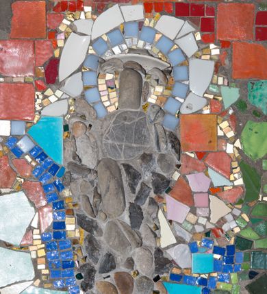 Zdjęcie nr 1: Mozaika w kształcie stojącego prostokąta z umieszczonym centralnie frontalnym przedstawieniem św. Józefa trzymającego w ramionach Dzieciątko Jezus. Józef ukazany całopostaciowo, w pozycji stojącej, na prawym ramieniu podtrzymuje Dzieciątko. Twarz świętego pozbawiona jest szczegółów fizjonomicznych. Wokół jego głowy znajduje się nimb w kolorach białym, szarym, złotym oraz jasnobłękitnym. Postać ubrana jest w ciemnoszare, długie szaty. Dzieciątko ukazane jest w dużym uproszczeniu, z pominięciem szczegółów fizjonomicznych i anatomicznych. Tło przedstawienia stanowi wielobarwna kompozycja z geometrycznych fragmentów ceramiki w różnych odcieniach czerwieni, zieleni, błękitu oraz granatu. 


