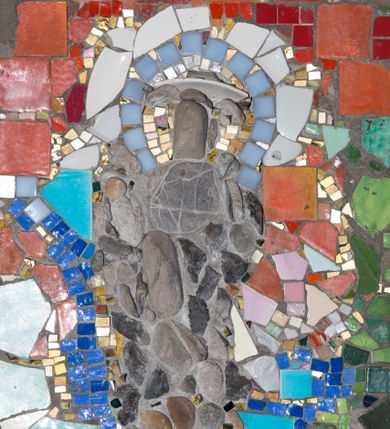 Zdjęcie nr 1: Mozaika w kształcie stojącego prostokąta z umieszczonym centralnie frontalnym przedstawieniem św. Józefa trzymającego w ramionach Dzieciątko Jezus. Józef ukazany całopostaciowo, w pozycji stojącej, na prawym ramieniu podtrzymuje Dzieciątko. Twarz świętego pozbawiona jest szczegółów fizjonomicznych. Wokół jego głowy znajduje się nimb w kolorach białym, szarym, złotym oraz jasnobłękitnym. Postać ubrana jest w ciemnoszare, długie szaty. Dzieciątko ukazane jest w dużym uproszczeniu, z pominięciem szczegółów fizjonomicznych i anatomicznych. Tło przedstawienia stanowi wielobarwna kompozycja z geometrycznych fragmentów ceramiki w różnych odcieniach czerwieni, zieleni, błękitu oraz granatu. 


