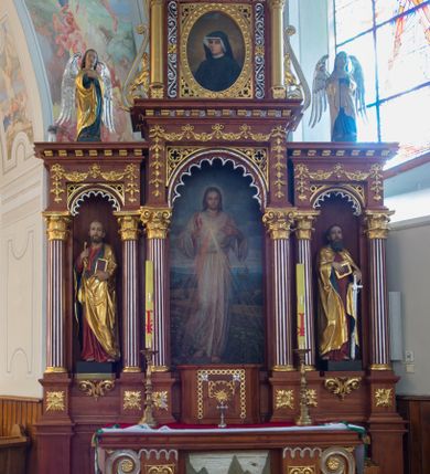 Zdjęcie nr 2: Przyścienną nastawę architektoniczną, trójosiową wykonano z drewna modrzewiowego. Jest to konstrukcja jednokondygnacyjna w formie łuku triumfalnego ze zwieńczeniem, ustawiona na dwustrefowym cokole. Stipes ołtarzowy uzyskał formę leżącego prostopadłościanu flankowanego po bokach spływami wolutowymi. W stipesie od przodu we wnęce pomiędzy dwoma pilastrami podtrzymującymi mensę umieszczono płaskorzeźbę z przedstawieniem Mojżesza wyprowadzającego wodę ze skały. Trzony pilastrów ozdobiono płaskorzeźbionym i złoconym ornamentem roślinnym. Na mensie na osi ustawiono
tabernakulum w formie leżącego prostopadłościanu o ściętych narożach, na niskim cokole, z profilowanym, niewielkim gzymsem, flankowanym na narożach pilastrami. Ścianka frontowa obwiedziona została rzędem złoconych okręgów. Na drzwiczkach umieszczono symbol Opatrzności Bożej od góry ujęte półkolistym łukiem. Oś środkową nastawy wydzielają dwie kolumny o kapitelach kompozytowych i kanelowanych trzonach dźwigających wydłużone imposty. Kolumny zostały połączone nad polem środkowym fryzem i gzymsem. Pole środkowe wypełnia nisza w formie stojącego prostokąta zamkniętego łukiem półkolistym z arkaturą z figurą św. Antoniego  Padwy oraz dwoma obrazami na zasuwach - "Niewiasty idące z Golgoty" oraz "Miłosierdzie Boże". W osiach zewnętrznych usytuowano niższe flankowane kanelowanymi kolumnami z kapitelami kompozytowymi, podtrzymującymi imposty połączone nad polem fryzem i gzymsem. Nisze uzyskały kształt stojącego prostokąta zamkniętego łukiem półkolistym z arkaturą z figurami apostołów św. Piotra i św. Pawła. Ponad niszami na belkowaniu usytuowano cokoły z figurami stojących aniołów ze złożonymi do modlitwy rękami. Zwieńczenie uzyskało kształt stojącego prostokąta flankowanego dwiema parami złoconych kolumienek, dźwigających imposty i gzyms nad polem środkowym wyłamanym w trójkąt. Po bokach widoczne są spływy wolutowe. Gzyms wieńczy krzyż i dwie złocone wazy. W polu zwieńczenia znajduje się rama w formie prostokąta z wpisanym owalem z obrazem św. Siostry Faustyny Kowalskiej. Ponad ramą widoczny jest chrystogram "IHS". Cokoły i imposty dekorowane zostały stylizowaną ornamentyką roślinną w postaci zwisów, kampanuli i girland. Struktura nastawy utrzymana jest w kolorze naturalnego drewna, natomiast ornamentyka, detale i profile zostały wyzłocone i wysrebrzone. Z kolei figury uzyskały naturalistyczną polichromię.