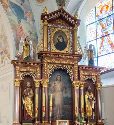 Zdjęcie nr 1: Przyścienną nastawę architektoniczną, trójosiową wykonano z drewna modrzewiowego. Jest to konstrukcja jednokondygnacyjna w formie łuku triumfalnego ze zwieńczeniem, ustawiona na dwustrefowym cokole. Stipes ołtarzowy uzyskał formę leżącego prostopadłościanu flankowanego po bokach spływami wolutowymi. W stipesie od przodu we wnęce pomiędzy dwoma pilastrami podtrzymującymi mensę umieszczono płaskorzeźbę z przedstawieniem Mojżesza wyprowadzającego wodę ze skały. Trzony pilastrów ozdobiono płaskorzeźbionym i złoconym ornamentem roślinnym. Na mensie na osi ustawiono
tabernakulum w formie leżącego prostopadłościanu o ściętych narożach, na niskim cokole, z profilowanym, niewielkim gzymsem, flankowanym na narożach pilastrami. Ścianka frontowa obwiedziona została rzędem złoconych okręgów. Na drzwiczkach umieszczono symbol Opatrzności Bożej od góry ujęte półkolistym łukiem. Oś środkową nastawy wydzielają dwie kolumny o kapitelach kompozytowych i kanelowanych trzonach dźwigających wydłużone imposty. Kolumny zostały połączone nad polem środkowym fryzem i gzymsem. Pole środkowe wypełnia nisza w formie stojącego prostokąta zamkniętego łukiem półkolistym z arkaturą z figurą św. Antoniego  Padwy oraz dwoma obrazami na zasuwach - "Niewiasty idące z Golgoty" oraz "Miłosierdzie Boże". W osiach zewnętrznych usytuowano niższe flankowane kanelowanymi kolumnami z kapitelami kompozytowymi, podtrzymującymi imposty połączone nad polem fryzem i gzymsem. Nisze uzyskały kształt stojącego prostokąta zamkniętego łukiem półkolistym z arkaturą z figurami apostołów św. Piotra i św. Pawła. Ponad niszami na belkowaniu usytuowano cokoły z figurami stojących aniołów ze złożonymi do modlitwy rękami. Zwieńczenie uzyskało kształt stojącego prostokąta flankowanego dwiema parami złoconych kolumienek, dźwigających imposty i gzyms nad polem środkowym wyłamanym w trójkąt. Po bokach widoczne są spływy wolutowe. Gzyms wieńczy krzyż i dwie złocone wazy. W polu zwieńczenia znajduje się rama w formie prostokąta z wpisanym owalem z obrazem św. Siostry Faustyny Kowalskiej. Ponad ramą widoczny jest chrystogram "IHS". Cokoły i imposty dekorowane zostały stylizowaną ornamentyką roślinną w postaci zwisów, kampanuli i girland. Struktura nastawy utrzymana jest w kolorze naturalnego drewna, natomiast ornamentyka, detale i profile zostały wyzłocone i wysrebrzone. Z kolei figury uzyskały naturalistyczną polichromię.