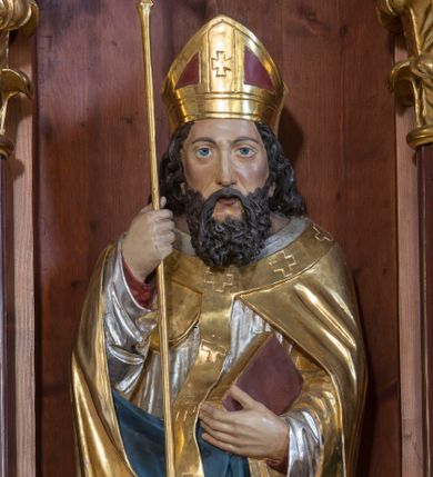 Zdjęcie nr 1: Rzeźba z przedstawieniem św. Wojciecha, biskupa jest drążona od tyłu. Biskup został ustawiony na prostopadłościennym postumencie. Świętego przedstawiono frontalnie w całej postaci w nieznacznym kontrapoście. W prawej ręce trzyma laskę krzyżową, a w lewej przyciśniętej do torsu zamknietą księgę, podtrzymując jednocześnie prawą połę kapy. Męczennika ukazano jako dojrzałego mężczyznę z pociągłą twarzą, długimi, ciemnymi, falowanymi włosami spływającymi na ramiona oraz brodą. Ma prosty, długi nos, oraz wydatne usta i szeroko otwarte oczy o spojrzeniu skierowanym przed siebie. Ubrany jest w długą czerwoną albę odsłaniającą czubki ciemnych butów, srebrną dalmatykę z długimi rękawami, zdobioną u dołu geometryczną ornamentyką oraz złotą kapę z błękitnawą podszewką. Na kapie ma nałożony długi, złoty paliusz dekorowany krzyżykami równoramiennymi. Na jego głowie widoczna jest czerwono-złota infuła.