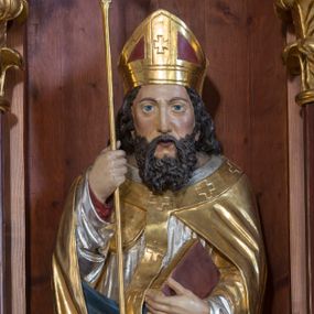 Zdjęcie nr 1: Rzeźba z przedstawieniem św. Wojciecha, biskupa jest drążona od tyłu. Biskup został ustawiony na prostopadłościennym postumencie. Świętego przedstawiono frontalnie w całej postaci w nieznacznym kontrapoście. W prawej ręce trzyma laskę krzyżową, a w lewej przyciśniętej do torsu zamknietą księgę, podtrzymując jednocześnie prawą połę kapy. Męczennika ukazano jako dojrzałego mężczyznę z pociągłą twarzą, długimi, ciemnymi, falowanymi włosami spływającymi na ramiona oraz brodą. Ma prosty, długi nos, oraz wydatne usta i szeroko otwarte oczy o spojrzeniu skierowanym przed siebie. Ubrany jest w długą czerwoną albę odsłaniającą czubki ciemnych butów, srebrną dalmatykę z długimi rękawami, zdobioną u dołu geometryczną ornamentyką oraz złotą kapę z błękitnawą podszewką. Na kapie ma nałożony długi, złoty paliusz dekorowany krzyżykami równoramiennymi. Na jego głowie widoczna jest czerwono-złota infuła.