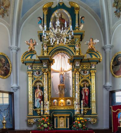 Zdjęcie nr 1: Ołtarz w kaplicy Matki Boskiej Szaflarskiej stanowi przykład przyściennej, architektonicznej, drewnianej nastawy trójosiowej ze zwieńczeniem. Stipes ołtarzowy z drewnianą mensą z kamiennym portatylem uzyskał kształt leżącego prostopadłościanu. Jego przód flankują hermy o trzonach dekorowanych cekinami. W polu zastosowano prostokątną płycinę ujętą kimationem. Na mensie ustawiono prostopadłościenne tabernakulum z wyodrębnionym cokołem i gzymsem koronującym wyłamanym na osi, podtrzymywanym przez dwa pilastry. Krawędzie tabernakulum zdobi imitacja boniowania. Drzwiczki ozdobiono stojącą, prostokątną płyciną, zwieńczoną trójkątnym przyczółkiem. W polu płyciny umieszczono stojący na obłokach płaskorzeźbiony i złocony kielich z hostią otoczoną promieniami. Jednokondygnacyjna nastawa wspiera się na prostokątnej predelli, przechodzącej pod osiami bocznymi w wolutowe konsole. Osie retabulum wydzielone zostały za pomocą skrajnych pilastrów oraz pary kolumn flankujących oś środkową. Podpory dźwigają pełne belkowanie wyłamane na ich osi, przerwane w partii pola środkowego oraz na osiach skrajnych przerwany półokrągły przyczółek. Wewnętrzna para kolumn dźwiga dodatkowo parę konsol podtrzymujących pełne belkowanie wyłamane na ich osi. Zwieńczenie uzyskało formę kwadratu zamkniętego szczytem z krzyżem, ujętego parą kolumienek na cokołach podtrzymujących skrajne woluty. Po zewnętrznych bokach zwieńczenia usytuowano postacie adorujących aniołów. W polu środkowym kondygnacji znalazła się nisza w formie stojącego prostokąta zamkniętego archiwoltą opadającą na belkowanie z konchą wydzieloną przez poziomy gzyms wsparty na dwóch pilastrach. W podniebiu konchy widoczna jest rzeźbiona i srebrzona gołębica Ducha Świętego. W niszy ustawiono figurę Matki Boskiej z Dzieciątkiem stojącą na wysokim czworobocznym postumencie. W osiach bocznych znalazły się nisze zamknięte konchą, u dołu podtrzymywane wspornikiem, z figurami św. Elżbiety Węgierskiej i św. Kazimierza Królewicza. W polu zwieńczenia umieszczono obraz w formie owalu ujęty ramą z przedstawieniem św. Maurycego. Ołtarz w całości pokryty został bogatą i zróżnicowaną dekoracją ornamentalną. Liśćmi akantu ozdobiono cokoły kolumn i wolut podwieszonych pod boczne osie. Kolumny w jednej czwartej wysokości obwiedzione zostały profilowaną listwą wypełnioną rautami. Ich dolne części ozdobiono uskrzydlonymi główkami aniołów z podwieszonymi festonami i girlandami. Z kolei w górnych partiach wykorzystano spiralne wici winnego grona. Dekoracyjnie artykułowano także pilastry, które w jednej czwartej przedzielono profilowanym gzymsem. Ich dolne części szczelnie wypełniono ornamentem okuciowym z rautem w środku, z kolei górne pokryto układającą się swobodnie i symetrycznie wstęgą z kaboszonami. Fryz belkowania wypełniony został wicią roślinną. Pola pomiędzy niszą a belkowaniem ozdobiono uskrzydlonymi główkami aniołów i podwieszonymi pod nimi girlandami. Ponadto uskrzydlone główki aniołów zdobią imposty podstawy zwieńczenia oraz klucz archiwolty ponad niszą główną. Kolumieńki zwieńczenia ozdobione zostały układającą się spiralnie wstęgą. Na dekoracje ornamentalną nastawy składają się również różne zdobienia profilów belkowania i zwieńczenia w postaci kostkowania, wolich oczu oraz pasów utworzonych z liści akantu.
