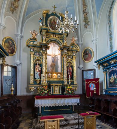 Zdjęcie nr 2: Ołtarz w kaplicy Matki Boskiej Szaflarskiej stanowi przykład przyściennej, architektonicznej, drewnianej nastawy trójosiowej ze zwieńczeniem. Stipes ołtarzowy z drewnianą mensą z kamiennym portatylem uzyskał kształt leżącego prostopadłościanu. Jego przód flankują hermy o trzonach dekorowanych cekinami. W polu zastosowano prostokątną płycinę ujętą kimationem. Na mensie ustawiono prostopadłościenne tabernakulum z wyodrębnionym cokołem i gzymsem koronującym wyłamanym na osi, podtrzymywanym przez dwa pilastry. Krawędzie tabernakulum zdobi imitacja boniowania. Drzwiczki ozdobiono stojącą, prostokątną płyciną, zwieńczoną trójkątnym przyczółkiem. W polu płyciny umieszczono stojący na obłokach płaskorzeźbiony i złocony kielich z hostią otoczoną promieniami. Jednokondygnacyjna nastawa wspiera się na prostokątnej predelli, przechodzącej pod osiami bocznymi w wolutowe konsole. Osie retabulum wydzielone zostały za pomocą skrajnych pilastrów oraz pary kolumn flankujących oś środkową. Podpory dźwigają pełne belkowanie wyłamane na ich osi, przerwane w partii pola środkowego oraz na osiach skrajnych przerwany półokrągły przyczółek. Wewnętrzna para kolumn dźwiga dodatkowo parę konsol podtrzymujących pełne belkowanie wyłamane na ich osi. Zwieńczenie uzyskało formę kwadratu zamkniętego szczytem z krzyżem, ujętego parą kolumienek na cokołach podtrzymujących skrajne woluty. Po zewnętrznych bokach zwieńczenia usytuowano postacie adorujących aniołów. W polu środkowym kondygnacji znalazła się nisza w formie stojącego prostokąta zamkniętego archiwoltą opadającą na belkowanie z konchą wydzieloną przez poziomy gzyms wsparty na dwóch pilastrach. W podniebiu konchy widoczna jest rzeźbiona i srebrzona gołębica Ducha Świętego. W niszy ustawiono figurę Matki Boskiej z Dzieciątkiem stojącą na wysokim czworobocznym postumencie. W osiach bocznych znalazły się nisze zamknięte konchą, u dołu podtrzymywane wspornikiem, z figurami św. Elżbiety Węgierskiej i św. Kazimierza Królewicza. W polu zwieńczenia umieszczono obraz w formie owalu ujęty ramą z przedstawieniem św. Maurycego. Ołtarz w całości pokryty został bogatą i zróżnicowaną dekoracją ornamentalną. Liśćmi akantu ozdobiono cokoły kolumn i wolut podwieszonych pod boczne osie. Kolumny w jednej czwartej wysokości obwiedzione zostały profilowaną listwą wypełnioną rautami. Ich dolne części ozdobiono uskrzydlonymi główkami aniołów z podwieszonymi festonami i girlandami. Z kolei w górnych partiach wykorzystano spiralne wici winnego grona. Dekoracyjnie artykułowano także pilastry, które w jednej czwartej przedzielono profilowanym gzymsem. Ich dolne części szczelnie wypełniono ornamentem okuciowym z rautem w środku, z kolei górne pokryto układającą się swobodnie i symetrycznie wstęgą z kaboszonami. Fryz belkowania wypełniony został wicią roślinną. Pola pomiędzy niszą a belkowaniem ozdobiono uskrzydlonymi główkami aniołów i podwieszonymi pod nimi girlandami. Ponadto uskrzydlone główki aniołów zdobią imposty podstawy zwieńczenia oraz klucz archiwolty ponad niszą główną. Kolumieńki zwieńczenia ozdobione zostały układającą się spiralnie wstęgą. Na dekoracje ornamentalną nastawy składają się również różne zdobienia profilów belkowania i zwieńczenia w postaci kostkowania, wolich oczu oraz pasów utworzonych z liści akantu.
