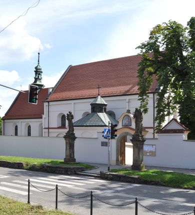 Zdjęcie nr 6: Kościół jest położony na wzgórzu w pobliżu centrum, przy ul. Brata Alojzego Kosiby. Od zachodu przed kościołem znajduje się dziedziniec i dawny cmentarz obwiedzione murem z bramkami i stacjami Męki Pańskiej. Z cmentarza wejście prowadzi do kościoła, a przez furtę na teren przyklasztorny i dalej do ogrodu klasztornego. Kościół wzniesiony jest z cegły, otynkowany. Świątynia jest orientowana, jednonawowa, z węższym prezbiterium, z kaplicą od północy i kruchtą od zachodu. Przy kościele od południa znajdują się zakrystia i zabudowania klasztorne. Prezbiterium na rzucie prostokąta, zamknięte od wschodu prostą ścianą, trójprzęsłowe, przekryte sklepieniem kolebkowym z przecinającymi się lunetami. W każdym przęśle ściany północnej wysoko umieszczone półkoliście zamknięte okna. Otworom okiennym odpowiadają tej samej wielkości nisze umieszczone w ścianie południowej. Wejście do klasztornego korytarza i zakrystii umieszczone jest w przęśle środkowym. Odpowiada mu otwór komunikacyjny po stronie przeciwnej. W przęśle zachodnim zlokalizowano drugie wejście do południowego korytarza klasztornego. Przestrzeń miedzy prezbiterium a korpusem zawężona arkadą tęczową, zamkniętą półkoliście. Korpus jednonawowy, szerszy z wnękami na ołtarze, czteroprzęsłowy, przekryty sklepieniem kolebkowym na gurtach z lunetami. Wnęka w trzecim przęśle od wschodu zamieniona na otwór do kaplicy północnej. Połowę przęsła zachodniego zajmuje empora muzyczna oparta na nisko schodzącym sklepieniu kolebkowym. Przy emporze, w ścianach wzdłużnych nawy, znacznie mniejsze, półkoliście zamknięte wnęki na konfesjonały. W lunetach, po obu stronach półkoliście zamknięte okna. Okna w przęśle zachodnim zastępują tej samej wielkości nisze. Główne wejście do kościoła w osi ściany zachodniej, zamknięte łukiem odcinkowym. W osi nad chórem muzycznym, półkoliście zamknięte okno, większe od pozostałych. Wejście do kościoła objęte prostokątnym portalem z piaskowca. Kaplica północna wzniesiona na planie kwadratu, przekryta sklepieniem współczesnym, zbliżonym w formie do klasztornego, zwieńczona latarnią. Okna, umieszczone w ścianie wschodniej i zachodniej, o tym samym wykroju utworzonym z wąskiego prostokąta przechodzącego górą w formę wachlarza lub wycinka koła. Zewnętrzne elewacje artykułuje podział osiowy i dekoracja lizenowo-ramowa, złożona z wydatnych elementów wertykalnych i horyzontalnych. Jednoosiową fasadę nad kruchtą ozdabia poniżej okna profilowany gzyms. Całość zamknięta wysoką, trójkątną ścianą szczytową, z trzema półkoliście zamkniętymi niszami. W nich polichromowane płaskorzeźby o kształcie stojącego prostokąta zamkniętego łukiem pełnym przedstawiające: stygmatyzację św. Franciszka, św. Piotra z Alkantary, św. Antoniego Padewskiego. Szczyt zwieńczony metalowym krzyżem osadzonym na niskim cokole. Kruchta przy fasadzie  z dwoma wejściami. Wejście zachodnie obejmuje półkoliście zamknięty portal z piaskowca, z dekoracją płycinową na węgarach, z kapitelami i skromnie profilowaną archiwoltą spiętą kluczem. Węższe wejście od północy z prostym, półkoliście zamkniętym portalem z piaskowca. Budowlę zamkniętą profilowanym gzymsem wieńczy trójkątny naczółek, przechodzący po bokach w balustradę z dekoracją płycinową. Na narożnych słupkach balustrady obeliskowe sterczyny. W półkoliście zamkniętej wnęce, umieszczonej pośrodku naczółka, wizerunek Matki Boskiej Nieustającej Pomocy. Wschodnia ściana prezbiterium, w dolnej partii zupełnie płaska, w górnej z triadą prostokątnych wnęk. Część szczytową oddziela od dolnej profilowany gzyms kryty dachówką. W osi trójkątnego szczytu wschodniego prostokątna wnęka z płaskorzeźbioną i polichromowaną sceną przedstawiającą św. Porcjunkulę w towarzystwie Chrystusa oraz św. Franciszka. Bryłę świątyni zamykają wysokie dachy dwuspadowe. Pośrodku dachu prezbiterium wieloboczna wieżyczka na sygnaturkę z ażurową latarnią. Bryłę kaplicy przekrywa dach czterospadowy. Ośmioboczną formę jej latarni zamyka dach wielospadowy zwieńczony metalowym krzyżem.