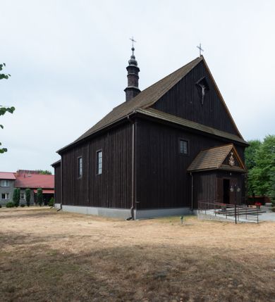 Zdjęcie nr 2: Kościół z drewna modrzewiowego o konstrukcji zrębowej, na betonowej podmurówce, otoczony drewnianym ogrodzeniem. Bryła składa się z korpusu nawowego na planie prostokąta oraz z wydłużonego, prostokątnego prezbiterium z zakrystią na przedłużeniu, węższego od nawy, zakończonego ścianą prostą, skierowanego na południe. Od strony północnej dobudowana jest prostokątna kruchta, od zachodu dwa przedsionki na dwóch skrajnych bokach –  przy nawie i prezbiterium. 
Trójprzęsłowe prezbiterium nakryte jest  sklepieniem pozornym zwierciadlanym, które przedłuża się nad nawę środkową. Ściany prezbiterium artykułowane pilastrami dzielą je na trzy przęsła. Kapitele pilastrów dźwigają nasadniki, które podtrzymują gzymsik. Przedłużeniem kapiteli w strefie stropu są płaskie belki biegnące przez całą jego szerokość, wyznaczając tym samym pola, w których umieszczono polichromię. Po obu stronach ołtarza drzwi w kształcie stojącego prostokąta. Prezbiterium od korpusu nawowego oddzielone silnie wystającą belką sklepienia oraz belką tęczową z grupą Ukrzyżowania. 
Korpus o trzech nawach o układzie halowym, z których środkowa jest dwa razy szersza od bocznych, trójprzęsłowa, z nawami oddzielonymi kanelowanymi filarami z nasadnikami podtrzymującymi gzyms. Nawy boczne nakryte kolebką. Na linii filarów pilastry na ścianach, łączą je poprzeczne belki sklepienia. W ostatnim przęśle chór muzyczny wsparty na dwóch filarach, z pełnym parapetem występującym w części środkowej trójbocznie do przodu. Jego krawędź dolna dekorowana jest lambrekinem. Ściany kościoła pokryte polichormią, pomalowane na jasnożółto, z jasnozielonymi podporami i brązowymi liniami podziału stropu. Te elementy wyznaczają ramy wypełnione polami z dekoracją ornamentalną w kształcie prostokątów i scenami figuralnymi zamkniętymi w kołach. 
Otwory okienne w drugim i trzecim przęśle nawy, od wschodu i zachodu, prostokątne. Także w prezbiterium po jednym prostokątnym oknie z każdej strony. Od strony wejścia dwa niewielkie okna prostokątne ponad kruchtą. Na ścianie zamknięcia zakrystii cztery prostokątne, symetrycznie rozłożone okna. 
 Fasada poprzedzona przedsionkiem w kształcie prostopadłościanu nakrytego dachem dwuspadowym, z przyczółkiem. Otwór drzwiowy w formie stojącego prostokąta, a w szczycie przyczółka trójkątne, wielodzielne okienko. Fasada w kształcie leżącego prostokąta z trójkątnym szczytem, części oddzielone daszkiem pulpitowym. Po dwóch stronach przedsionka prostokątne okna, w szczycie fasady  nakryty daszkiem krucyfiks. 
Elewacje zewnętrzne na podmurówce, artykułowane jedynie pionowymi, wystającymi przed lico belkami, przeprute oknami. Nad nawą i prezbiterium dachy dwuspadowe, nad prezbiterium niższy niż nad nawą, kryty blachą. Nad nawą wieżyczka na sygnaturkę zwieńczona cebulastym hełmem i krzyżem na szczycie. Na końcach dachu przy prezbiterium i nawie metalowe krzyże. 
