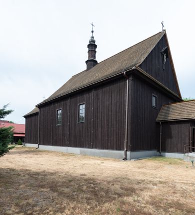 Zdjęcie nr 13: Kościół z drewna modrzewiowego o konstrukcji zrębowej, na betonowej podmurówce, otoczony drewnianym ogrodzeniem. Bryła składa się z korpusu nawowego na planie prostokąta oraz z wydłużonego, prostokątnego prezbiterium z zakrystią na przedłużeniu, węższego od nawy, zakończonego ścianą prostą, skierowanego na południe. Od strony północnej dobudowana jest prostokątna kruchta, od zachodu dwa przedsionki na dwóch skrajnych bokach –  przy nawie i prezbiterium. 
Trójprzęsłowe prezbiterium nakryte jest  sklepieniem pozornym zwierciadlanym, które przedłuża się nad nawę środkową. Ściany prezbiterium artykułowane pilastrami dzielą je na trzy przęsła. Kapitele pilastrów dźwigają nasadniki, które podtrzymują gzymsik. Przedłużeniem kapiteli w strefie stropu są płaskie belki biegnące przez całą jego szerokość, wyznaczając tym samym pola, w których umieszczono polichromię. Po obu stronach ołtarza drzwi w kształcie stojącego prostokąta. Prezbiterium od korpusu nawowego oddzielone silnie wystającą belką sklepienia oraz belką tęczową z grupą Ukrzyżowania. 
Korpus o trzech nawach o układzie halowym, z których środkowa jest dwa razy szersza od bocznych, trójprzęsłowa, z nawami oddzielonymi kanelowanymi filarami z nasadnikami podtrzymującymi gzyms. Nawy boczne nakryte kolebką. Na linii filarów pilastry na ścianach, łączą je poprzeczne belki sklepienia. W ostatnim przęśle chór muzyczny wsparty na dwóch filarach, z pełnym parapetem występującym w części środkowej trójbocznie do przodu. Jego krawędź dolna dekorowana jest lambrekinem. Ściany kościoła pokryte polichormią, pomalowane na jasnożółto, z jasnozielonymi podporami i brązowymi liniami podziału stropu. Te elementy wyznaczają ramy wypełnione polami z dekoracją ornamentalną w kształcie prostokątów i scenami figuralnymi zamkniętymi w kołach. 
Otwory okienne w drugim i trzecim przęśle nawy, od wschodu i zachodu, prostokątne. Także w prezbiterium po jednym prostokątnym oknie z każdej strony. Od strony wejścia dwa niewielkie okna prostokątne ponad kruchtą. Na ścianie zamknięcia zakrystii cztery prostokątne, symetrycznie rozłożone okna. 
 Fasada poprzedzona przedsionkiem w kształcie prostopadłościanu nakrytego dachem dwuspadowym, z przyczółkiem. Otwór drzwiowy w formie stojącego prostokąta, a w szczycie przyczółka trójkątne, wielodzielne okienko. Fasada w kształcie leżącego prostokąta z trójkątnym szczytem, części oddzielone daszkiem pulpitowym. Po dwóch stronach przedsionka prostokątne okna, w szczycie fasady  nakryty daszkiem krucyfiks. 
Elewacje zewnętrzne na podmurówce, artykułowane jedynie pionowymi, wystającymi przed lico belkami, przeprute oknami. Nad nawą i prezbiterium dachy dwuspadowe, nad prezbiterium niższy niż nad nawą, kryty blachą. Nad nawą wieżyczka na sygnaturkę zwieńczona cebulastym hełmem i krzyżem na szczycie. Na końcach dachu przy prezbiterium i nawie metalowe krzyże. 
