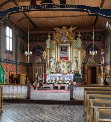 Zdjęcie nr 14: Kościół z drewna modrzewiowego o konstrukcji zrębowej, na betonowej podmurówce, otoczony drewnianym ogrodzeniem. Bryła składa się z korpusu nawowego na planie prostokąta oraz z wydłużonego, prostokątnego prezbiterium z zakrystią na przedłużeniu, węższego od nawy, zakończonego ścianą prostą, skierowanego na południe. Od strony północnej dobudowana jest prostokątna kruchta, od zachodu dwa przedsionki na dwóch skrajnych bokach –  przy nawie i prezbiterium. 
Trójprzęsłowe prezbiterium nakryte jest  sklepieniem pozornym zwierciadlanym, które przedłuża się nad nawę środkową. Ściany prezbiterium artykułowane pilastrami dzielą je na trzy przęsła. Kapitele pilastrów dźwigają nasadniki, które podtrzymują gzymsik. Przedłużeniem kapiteli w strefie stropu są płaskie belki biegnące przez całą jego szerokość, wyznaczając tym samym pola, w których umieszczono polichromię. Po obu stronach ołtarza drzwi w kształcie stojącego prostokąta. Prezbiterium od korpusu nawowego oddzielone silnie wystającą belką sklepienia oraz belką tęczową z grupą Ukrzyżowania. 
Korpus o trzech nawach o układzie halowym, z których środkowa jest dwa razy szersza od bocznych, trójprzęsłowa, z nawami oddzielonymi kanelowanymi filarami z nasadnikami podtrzymującymi gzyms. Nawy boczne nakryte kolebką. Na linii filarów pilastry na ścianach, łączą je poprzeczne belki sklepienia. W ostatnim przęśle chór muzyczny wsparty na dwóch filarach, z pełnym parapetem występującym w części środkowej trójbocznie do przodu. Jego krawędź dolna dekorowana jest lambrekinem. Ściany kościoła pokryte polichormią, pomalowane na jasnożółto, z jasnozielonymi podporami i brązowymi liniami podziału stropu. Te elementy wyznaczają ramy wypełnione polami z dekoracją ornamentalną w kształcie prostokątów i scenami figuralnymi zamkniętymi w kołach. 
Otwory okienne w drugim i trzecim przęśle nawy, od wschodu i zachodu, prostokątne. Także w prezbiterium po jednym prostokątnym oknie z każdej strony. Od strony wejścia dwa niewielkie okna prostokątne ponad kruchtą. Na ścianie zamknięcia zakrystii cztery prostokątne, symetrycznie rozłożone okna. 
 Fasada poprzedzona przedsionkiem w kształcie prostopadłościanu nakrytego dachem dwuspadowym, z przyczółkiem. Otwór drzwiowy w formie stojącego prostokąta, a w szczycie przyczółka trójkątne, wielodzielne okienko. Fasada w kształcie leżącego prostokąta z trójkątnym szczytem, części oddzielone daszkiem pulpitowym. Po dwóch stronach przedsionka prostokątne okna, w szczycie fasady  nakryty daszkiem krucyfiks. 
Elewacje zewnętrzne na podmurówce, artykułowane jedynie pionowymi, wystającymi przed lico belkami, przeprute oknami. Nad nawą i prezbiterium dachy dwuspadowe, nad prezbiterium niższy niż nad nawą, kryty blachą. Nad nawą wieżyczka na sygnaturkę zwieńczona cebulastym hełmem i krzyżem na szczycie. Na końcach dachu przy prezbiterium i nawie metalowe krzyże. 
