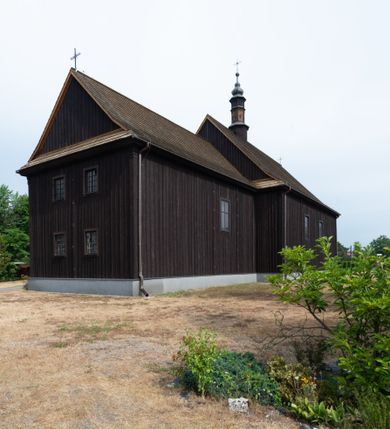 Zdjęcie nr 24: Kościół z drewna modrzewiowego o konstrukcji zrębowej, na betonowej podmurówce, otoczony drewnianym ogrodzeniem. Bryła składa się z korpusu nawowego na planie prostokąta oraz z wydłużonego, prostokątnego prezbiterium z zakrystią na przedłużeniu, węższego od nawy, zakończonego ścianą prostą, skierowanego na południe. Od strony północnej dobudowana jest prostokątna kruchta, od zachodu dwa przedsionki na dwóch skrajnych bokach –  przy nawie i prezbiterium. 
Trójprzęsłowe prezbiterium nakryte jest  sklepieniem pozornym zwierciadlanym, które przedłuża się nad nawę środkową. Ściany prezbiterium artykułowane pilastrami dzielą je na trzy przęsła. Kapitele pilastrów dźwigają nasadniki, które podtrzymują gzymsik. Przedłużeniem kapiteli w strefie stropu są płaskie belki biegnące przez całą jego szerokość, wyznaczając tym samym pola, w których umieszczono polichromię. Po obu stronach ołtarza drzwi w kształcie stojącego prostokąta. Prezbiterium od korpusu nawowego oddzielone silnie wystającą belką sklepienia oraz belką tęczową z grupą Ukrzyżowania. 
Korpus o trzech nawach o układzie halowym, z których środkowa jest dwa razy szersza od bocznych, trójprzęsłowa, z nawami oddzielonymi kanelowanymi filarami z nasadnikami podtrzymującymi gzyms. Nawy boczne nakryte kolebką. Na linii filarów pilastry na ścianach, łączą je poprzeczne belki sklepienia. W ostatnim przęśle chór muzyczny wsparty na dwóch filarach, z pełnym parapetem występującym w części środkowej trójbocznie do przodu. Jego krawędź dolna dekorowana jest lambrekinem. Ściany kościoła pokryte polichormią, pomalowane na jasnożółto, z jasnozielonymi podporami i brązowymi liniami podziału stropu. Te elementy wyznaczają ramy wypełnione polami z dekoracją ornamentalną w kształcie prostokątów i scenami figuralnymi zamkniętymi w kołach. 
Otwory okienne w drugim i trzecim przęśle nawy, od wschodu i zachodu, prostokątne. Także w prezbiterium po jednym prostokątnym oknie z każdej strony. Od strony wejścia dwa niewielkie okna prostokątne ponad kruchtą. Na ścianie zamknięcia zakrystii cztery prostokątne, symetrycznie rozłożone okna. 
 Fasada poprzedzona przedsionkiem w kształcie prostopadłościanu nakrytego dachem dwuspadowym, z przyczółkiem. Otwór drzwiowy w formie stojącego prostokąta, a w szczycie przyczółka trójkątne, wielodzielne okienko. Fasada w kształcie leżącego prostokąta z trójkątnym szczytem, części oddzielone daszkiem pulpitowym. Po dwóch stronach przedsionka prostokątne okna, w szczycie fasady  nakryty daszkiem krucyfiks. 
Elewacje zewnętrzne na podmurówce, artykułowane jedynie pionowymi, wystającymi przed lico belkami, przeprute oknami. Nad nawą i prezbiterium dachy dwuspadowe, nad prezbiterium niższy niż nad nawą, kryty blachą. Nad nawą wieżyczka na sygnaturkę zwieńczona cebulastym hełmem i krzyżem na szczycie. Na końcach dachu przy prezbiterium i nawie metalowe krzyże. 

