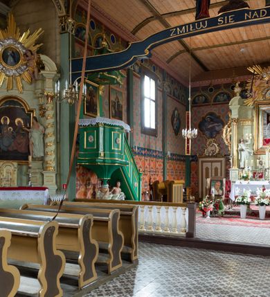 Zdjęcie nr 15: Kościół z drewna modrzewiowego o konstrukcji zrębowej, na betonowej podmurówce, otoczony drewnianym ogrodzeniem. Bryła składa się z korpusu nawowego na planie prostokąta oraz z wydłużonego, prostokątnego prezbiterium z zakrystią na przedłużeniu, węższego od nawy, zakończonego ścianą prostą, skierowanego na południe. Od strony północnej dobudowana jest prostokątna kruchta, od zachodu dwa przedsionki na dwóch skrajnych bokach –  przy nawie i prezbiterium. 
Trójprzęsłowe prezbiterium nakryte jest  sklepieniem pozornym zwierciadlanym, które przedłuża się nad nawę środkową. Ściany prezbiterium artykułowane pilastrami dzielą je na trzy przęsła. Kapitele pilastrów dźwigają nasadniki, które podtrzymują gzymsik. Przedłużeniem kapiteli w strefie stropu są płaskie belki biegnące przez całą jego szerokość, wyznaczając tym samym pola, w których umieszczono polichromię. Po obu stronach ołtarza drzwi w kształcie stojącego prostokąta. Prezbiterium od korpusu nawowego oddzielone silnie wystającą belką sklepienia oraz belką tęczową z grupą Ukrzyżowania. 
Korpus o trzech nawach o układzie halowym, z których środkowa jest dwa razy szersza od bocznych, trójprzęsłowa, z nawami oddzielonymi kanelowanymi filarami z nasadnikami podtrzymującymi gzyms. Nawy boczne nakryte kolebką. Na linii filarów pilastry na ścianach, łączą je poprzeczne belki sklepienia. W ostatnim przęśle chór muzyczny wsparty na dwóch filarach, z pełnym parapetem występującym w części środkowej trójbocznie do przodu. Jego krawędź dolna dekorowana jest lambrekinem. Ściany kościoła pokryte polichormią, pomalowane na jasnożółto, z jasnozielonymi podporami i brązowymi liniami podziału stropu. Te elementy wyznaczają ramy wypełnione polami z dekoracją ornamentalną w kształcie prostokątów i scenami figuralnymi zamkniętymi w kołach. 
Otwory okienne w drugim i trzecim przęśle nawy, od wschodu i zachodu, prostokątne. Także w prezbiterium po jednym prostokątnym oknie z każdej strony. Od strony wejścia dwa niewielkie okna prostokątne ponad kruchtą. Na ścianie zamknięcia zakrystii cztery prostokątne, symetrycznie rozłożone okna. 
 Fasada poprzedzona przedsionkiem w kształcie prostopadłościanu nakrytego dachem dwuspadowym, z przyczółkiem. Otwór drzwiowy w formie stojącego prostokąta, a w szczycie przyczółka trójkątne, wielodzielne okienko. Fasada w kształcie leżącego prostokąta z trójkątnym szczytem, części oddzielone daszkiem pulpitowym. Po dwóch stronach przedsionka prostokątne okna, w szczycie fasady  nakryty daszkiem krucyfiks. 
Elewacje zewnętrzne na podmurówce, artykułowane jedynie pionowymi, wystającymi przed lico belkami, przeprute oknami. Nad nawą i prezbiterium dachy dwuspadowe, nad prezbiterium niższy niż nad nawą, kryty blachą. Nad nawą wieżyczka na sygnaturkę zwieńczona cebulastym hełmem i krzyżem na szczycie. Na końcach dachu przy prezbiterium i nawie metalowe krzyże. 
