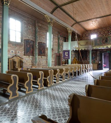 Zdjęcie nr 18: Kościół z drewna modrzewiowego o konstrukcji zrębowej, na betonowej podmurówce, otoczony drewnianym ogrodzeniem. Bryła składa się z korpusu nawowego na planie prostokąta oraz z wydłużonego, prostokątnego prezbiterium z zakrystią na przedłużeniu, węższego od nawy, zakończonego ścianą prostą, skierowanego na południe. Od strony północnej dobudowana jest prostokątna kruchta, od zachodu dwa przedsionki na dwóch skrajnych bokach –  przy nawie i prezbiterium. 
Trójprzęsłowe prezbiterium nakryte jest  sklepieniem pozornym zwierciadlanym, które przedłuża się nad nawę środkową. Ściany prezbiterium artykułowane pilastrami dzielą je na trzy przęsła. Kapitele pilastrów dźwigają nasadniki, które podtrzymują gzymsik. Przedłużeniem kapiteli w strefie stropu są płaskie belki biegnące przez całą jego szerokość, wyznaczając tym samym pola, w których umieszczono polichromię. Po obu stronach ołtarza drzwi w kształcie stojącego prostokąta. Prezbiterium od korpusu nawowego oddzielone silnie wystającą belką sklepienia oraz belką tęczową z grupą Ukrzyżowania. 
Korpus o trzech nawach o układzie halowym, z których środkowa jest dwa razy szersza od bocznych, trójprzęsłowa, z nawami oddzielonymi kanelowanymi filarami z nasadnikami podtrzymującymi gzyms. Nawy boczne nakryte kolebką. Na linii filarów pilastry na ścianach, łączą je poprzeczne belki sklepienia. W ostatnim przęśle chór muzyczny wsparty na dwóch filarach, z pełnym parapetem występującym w części środkowej trójbocznie do przodu. Jego krawędź dolna dekorowana jest lambrekinem. Ściany kościoła pokryte polichormią, pomalowane na jasnożółto, z jasnozielonymi podporami i brązowymi liniami podziału stropu. Te elementy wyznaczają ramy wypełnione polami z dekoracją ornamentalną w kształcie prostokątów i scenami figuralnymi zamkniętymi w kołach. 
Otwory okienne w drugim i trzecim przęśle nawy, od wschodu i zachodu, prostokątne. Także w prezbiterium po jednym prostokątnym oknie z każdej strony. Od strony wejścia dwa niewielkie okna prostokątne ponad kruchtą. Na ścianie zamknięcia zakrystii cztery prostokątne, symetrycznie rozłożone okna. 
 Fasada poprzedzona przedsionkiem w kształcie prostopadłościanu nakrytego dachem dwuspadowym, z przyczółkiem. Otwór drzwiowy w formie stojącego prostokąta, a w szczycie przyczółka trójkątne, wielodzielne okienko. Fasada w kształcie leżącego prostokąta z trójkątnym szczytem, części oddzielone daszkiem pulpitowym. Po dwóch stronach przedsionka prostokątne okna, w szczycie fasady  nakryty daszkiem krucyfiks. 
Elewacje zewnętrzne na podmurówce, artykułowane jedynie pionowymi, wystającymi przed lico belkami, przeprute oknami. Nad nawą i prezbiterium dachy dwuspadowe, nad prezbiterium niższy niż nad nawą, kryty blachą. Nad nawą wieżyczka na sygnaturkę zwieńczona cebulastym hełmem i krzyżem na szczycie. Na końcach dachu przy prezbiterium i nawie metalowe krzyże. 
