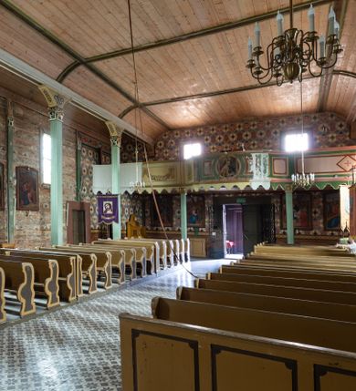 Zdjęcie nr 21: Kościół z drewna modrzewiowego o konstrukcji zrębowej, na betonowej podmurówce, otoczony drewnianym ogrodzeniem. Bryła składa się z korpusu nawowego na planie prostokąta oraz z wydłużonego, prostokątnego prezbiterium z zakrystią na przedłużeniu, węższego od nawy, zakończonego ścianą prostą, skierowanego na południe. Od strony północnej dobudowana jest prostokątna kruchta, od zachodu dwa przedsionki na dwóch skrajnych bokach –  przy nawie i prezbiterium. 
Trójprzęsłowe prezbiterium nakryte jest  sklepieniem pozornym zwierciadlanym, które przedłuża się nad nawę środkową. Ściany prezbiterium artykułowane pilastrami dzielą je na trzy przęsła. Kapitele pilastrów dźwigają nasadniki, które podtrzymują gzymsik. Przedłużeniem kapiteli w strefie stropu są płaskie belki biegnące przez całą jego szerokość, wyznaczając tym samym pola, w których umieszczono polichromię. Po obu stronach ołtarza drzwi w kształcie stojącego prostokąta. Prezbiterium od korpusu nawowego oddzielone silnie wystającą belką sklepienia oraz belką tęczową z grupą Ukrzyżowania. 
Korpus o trzech nawach o układzie halowym, z których środkowa jest dwa razy szersza od bocznych, trójprzęsłowa, z nawami oddzielonymi kanelowanymi filarami z nasadnikami podtrzymującymi gzyms. Nawy boczne nakryte kolebką. Na linii filarów pilastry na ścianach, łączą je poprzeczne belki sklepienia. W ostatnim przęśle chór muzyczny wsparty na dwóch filarach, z pełnym parapetem występującym w części środkowej trójbocznie do przodu. Jego krawędź dolna dekorowana jest lambrekinem. Ściany kościoła pokryte polichormią, pomalowane na jasnożółto, z jasnozielonymi podporami i brązowymi liniami podziału stropu. Te elementy wyznaczają ramy wypełnione polami z dekoracją ornamentalną w kształcie prostokątów i scenami figuralnymi zamkniętymi w kołach. 
Otwory okienne w drugim i trzecim przęśle nawy, od wschodu i zachodu, prostokątne. Także w prezbiterium po jednym prostokątnym oknie z każdej strony. Od strony wejścia dwa niewielkie okna prostokątne ponad kruchtą. Na ścianie zamknięcia zakrystii cztery prostokątne, symetrycznie rozłożone okna. 
 Fasada poprzedzona przedsionkiem w kształcie prostopadłościanu nakrytego dachem dwuspadowym, z przyczółkiem. Otwór drzwiowy w formie stojącego prostokąta, a w szczycie przyczółka trójkątne, wielodzielne okienko. Fasada w kształcie leżącego prostokąta z trójkątnym szczytem, części oddzielone daszkiem pulpitowym. Po dwóch stronach przedsionka prostokątne okna, w szczycie fasady  nakryty daszkiem krucyfiks. 
Elewacje zewnętrzne na podmurówce, artykułowane jedynie pionowymi, wystającymi przed lico belkami, przeprute oknami. Nad nawą i prezbiterium dachy dwuspadowe, nad prezbiterium niższy niż nad nawą, kryty blachą. Nad nawą wieżyczka na sygnaturkę zwieńczona cebulastym hełmem i krzyżem na szczycie. Na końcach dachu przy prezbiterium i nawie metalowe krzyże. 
