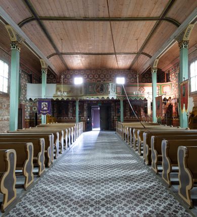 Zdjęcie nr 16: Kościół z drewna modrzewiowego o konstrukcji zrębowej, na betonowej podmurówce, otoczony drewnianym ogrodzeniem. Bryła składa się z korpusu nawowego na planie prostokąta oraz z wydłużonego, prostokątnego prezbiterium z zakrystią na przedłużeniu, węższego od nawy, zakończonego ścianą prostą, skierowanego na południe. Od strony północnej dobudowana jest prostokątna kruchta, od zachodu dwa przedsionki na dwóch skrajnych bokach –  przy nawie i prezbiterium. 
Trójprzęsłowe prezbiterium nakryte jest  sklepieniem pozornym zwierciadlanym, które przedłuża się nad nawę środkową. Ściany prezbiterium artykułowane pilastrami dzielą je na trzy przęsła. Kapitele pilastrów dźwigają nasadniki, które podtrzymują gzymsik. Przedłużeniem kapiteli w strefie stropu są płaskie belki biegnące przez całą jego szerokość, wyznaczając tym samym pola, w których umieszczono polichromię. Po obu stronach ołtarza drzwi w kształcie stojącego prostokąta. Prezbiterium od korpusu nawowego oddzielone silnie wystającą belką sklepienia oraz belką tęczową z grupą Ukrzyżowania. 
Korpus o trzech nawach o układzie halowym, z których środkowa jest dwa razy szersza od bocznych, trójprzęsłowa, z nawami oddzielonymi kanelowanymi filarami z nasadnikami podtrzymującymi gzyms. Nawy boczne nakryte kolebką. Na linii filarów pilastry na ścianach, łączą je poprzeczne belki sklepienia. W ostatnim przęśle chór muzyczny wsparty na dwóch filarach, z pełnym parapetem występującym w części środkowej trójbocznie do przodu. Jego krawędź dolna dekorowana jest lambrekinem. Ściany kościoła pokryte polichormią, pomalowane na jasnożółto, z jasnozielonymi podporami i brązowymi liniami podziału stropu. Te elementy wyznaczają ramy wypełnione polami z dekoracją ornamentalną w kształcie prostokątów i scenami figuralnymi zamkniętymi w kołach. 
Otwory okienne w drugim i trzecim przęśle nawy, od wschodu i zachodu, prostokątne. Także w prezbiterium po jednym prostokątnym oknie z każdej strony. Od strony wejścia dwa niewielkie okna prostokątne ponad kruchtą. Na ścianie zamknięcia zakrystii cztery prostokątne, symetrycznie rozłożone okna. 
 Fasada poprzedzona przedsionkiem w kształcie prostopadłościanu nakrytego dachem dwuspadowym, z przyczółkiem. Otwór drzwiowy w formie stojącego prostokąta, a w szczycie przyczółka trójkątne, wielodzielne okienko. Fasada w kształcie leżącego prostokąta z trójkątnym szczytem, części oddzielone daszkiem pulpitowym. Po dwóch stronach przedsionka prostokątne okna, w szczycie fasady  nakryty daszkiem krucyfiks. 
Elewacje zewnętrzne na podmurówce, artykułowane jedynie pionowymi, wystającymi przed lico belkami, przeprute oknami. Nad nawą i prezbiterium dachy dwuspadowe, nad prezbiterium niższy niż nad nawą, kryty blachą. Nad nawą wieżyczka na sygnaturkę zwieńczona cebulastym hełmem i krzyżem na szczycie. Na końcach dachu przy prezbiterium i nawie metalowe krzyże. 
