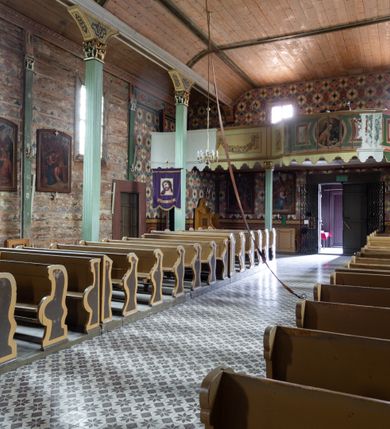 Zdjęcie nr 20: Kościół z drewna modrzewiowego o konstrukcji zrębowej, na betonowej podmurówce, otoczony drewnianym ogrodzeniem. Bryła składa się z korpusu nawowego na planie prostokąta oraz z wydłużonego, prostokątnego prezbiterium z zakrystią na przedłużeniu, węższego od nawy, zakończonego ścianą prostą, skierowanego na południe. Od strony północnej dobudowana jest prostokątna kruchta, od zachodu dwa przedsionki na dwóch skrajnych bokach –  przy nawie i prezbiterium. 
Trójprzęsłowe prezbiterium nakryte jest  sklepieniem pozornym zwierciadlanym, które przedłuża się nad nawę środkową. Ściany prezbiterium artykułowane pilastrami dzielą je na trzy przęsła. Kapitele pilastrów dźwigają nasadniki, które podtrzymują gzymsik. Przedłużeniem kapiteli w strefie stropu są płaskie belki biegnące przez całą jego szerokość, wyznaczając tym samym pola, w których umieszczono polichromię. Po obu stronach ołtarza drzwi w kształcie stojącego prostokąta. Prezbiterium od korpusu nawowego oddzielone silnie wystającą belką sklepienia oraz belką tęczową z grupą Ukrzyżowania. 
Korpus o trzech nawach o układzie halowym, z których środkowa jest dwa razy szersza od bocznych, trójprzęsłowa, z nawami oddzielonymi kanelowanymi filarami z nasadnikami podtrzymującymi gzyms. Nawy boczne nakryte kolebką. Na linii filarów pilastry na ścianach, łączą je poprzeczne belki sklepienia. W ostatnim przęśle chór muzyczny wsparty na dwóch filarach, z pełnym parapetem występującym w części środkowej trójbocznie do przodu. Jego krawędź dolna dekorowana jest lambrekinem. Ściany kościoła pokryte polichormią, pomalowane na jasnożółto, z jasnozielonymi podporami i brązowymi liniami podziału stropu. Te elementy wyznaczają ramy wypełnione polami z dekoracją ornamentalną w kształcie prostokątów i scenami figuralnymi zamkniętymi w kołach. 
Otwory okienne w drugim i trzecim przęśle nawy, od wschodu i zachodu, prostokątne. Także w prezbiterium po jednym prostokątnym oknie z każdej strony. Od strony wejścia dwa niewielkie okna prostokątne ponad kruchtą. Na ścianie zamknięcia zakrystii cztery prostokątne, symetrycznie rozłożone okna. 
 Fasada poprzedzona przedsionkiem w kształcie prostopadłościanu nakrytego dachem dwuspadowym, z przyczółkiem. Otwór drzwiowy w formie stojącego prostokąta, a w szczycie przyczółka trójkątne, wielodzielne okienko. Fasada w kształcie leżącego prostokąta z trójkątnym szczytem, części oddzielone daszkiem pulpitowym. Po dwóch stronach przedsionka prostokątne okna, w szczycie fasady  nakryty daszkiem krucyfiks. 
Elewacje zewnętrzne na podmurówce, artykułowane jedynie pionowymi, wystającymi przed lico belkami, przeprute oknami. Nad nawą i prezbiterium dachy dwuspadowe, nad prezbiterium niższy niż nad nawą, kryty blachą. Nad nawą wieżyczka na sygnaturkę zwieńczona cebulastym hełmem i krzyżem na szczycie. Na końcach dachu przy prezbiterium i nawie metalowe krzyże. 
