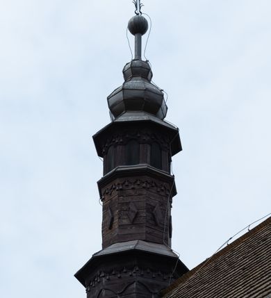 Zdjęcie nr 5: Kościół z drewna modrzewiowego o konstrukcji zrębowej, na betonowej podmurówce, otoczony drewnianym ogrodzeniem. Bryła składa się z korpusu nawowego na planie prostokąta oraz z wydłużonego, prostokątnego prezbiterium z zakrystią na przedłużeniu, węższego od nawy, zakończonego ścianą prostą, skierowanego na południe. Od strony północnej dobudowana jest prostokątna kruchta, od zachodu dwa przedsionki na dwóch skrajnych bokach –  przy nawie i prezbiterium. 
Trójprzęsłowe prezbiterium nakryte jest  sklepieniem pozornym zwierciadlanym, które przedłuża się nad nawę środkową. Ściany prezbiterium artykułowane pilastrami dzielą je na trzy przęsła. Kapitele pilastrów dźwigają nasadniki, które podtrzymują gzymsik. Przedłużeniem kapiteli w strefie stropu są płaskie belki biegnące przez całą jego szerokość, wyznaczając tym samym pola, w których umieszczono polichromię. Po obu stronach ołtarza drzwi w kształcie stojącego prostokąta. Prezbiterium od korpusu nawowego oddzielone silnie wystającą belką sklepienia oraz belką tęczową z grupą Ukrzyżowania. 
Korpus o trzech nawach o układzie halowym, z których środkowa jest dwa razy szersza od bocznych, trójprzęsłowa, z nawami oddzielonymi kanelowanymi filarami z nasadnikami podtrzymującymi gzyms. Nawy boczne nakryte kolebką. Na linii filarów pilastry na ścianach, łączą je poprzeczne belki sklepienia. W ostatnim przęśle chór muzyczny wsparty na dwóch filarach, z pełnym parapetem występującym w części środkowej trójbocznie do przodu. Jego krawędź dolna dekorowana jest lambrekinem. Ściany kościoła pokryte polichormią, pomalowane na jasnożółto, z jasnozielonymi podporami i brązowymi liniami podziału stropu. Te elementy wyznaczają ramy wypełnione polami z dekoracją ornamentalną w kształcie prostokątów i scenami figuralnymi zamkniętymi w kołach. 
Otwory okienne w drugim i trzecim przęśle nawy, od wschodu i zachodu, prostokątne. Także w prezbiterium po jednym prostokątnym oknie z każdej strony. Od strony wejścia dwa niewielkie okna prostokątne ponad kruchtą. Na ścianie zamknięcia zakrystii cztery prostokątne, symetrycznie rozłożone okna. 
 Fasada poprzedzona przedsionkiem w kształcie prostopadłościanu nakrytego dachem dwuspadowym, z przyczółkiem. Otwór drzwiowy w formie stojącego prostokąta, a w szczycie przyczółka trójkątne, wielodzielne okienko. Fasada w kształcie leżącego prostokąta z trójkątnym szczytem, części oddzielone daszkiem pulpitowym. Po dwóch stronach przedsionka prostokątne okna, w szczycie fasady  nakryty daszkiem krucyfiks. 
Elewacje zewnętrzne na podmurówce, artykułowane jedynie pionowymi, wystającymi przed lico belkami, przeprute oknami. Nad nawą i prezbiterium dachy dwuspadowe, nad prezbiterium niższy niż nad nawą, kryty blachą. Nad nawą wieżyczka na sygnaturkę zwieńczona cebulastym hełmem i krzyżem na szczycie. Na końcach dachu przy prezbiterium i nawie metalowe krzyże. 
