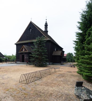 Zdjęcie nr 23: Kościół z drewna modrzewiowego o konstrukcji zrębowej, na betonowej podmurówce, otoczony drewnianym ogrodzeniem. Bryła składa się z korpusu nawowego na planie prostokąta oraz z wydłużonego, prostokątnego prezbiterium z zakrystią na przedłużeniu, węższego od nawy, zakończonego ścianą prostą, skierowanego na południe. Od strony północnej dobudowana jest prostokątna kruchta, od zachodu dwa przedsionki na dwóch skrajnych bokach –  przy nawie i prezbiterium. 
Trójprzęsłowe prezbiterium nakryte jest  sklepieniem pozornym zwierciadlanym, które przedłuża się nad nawę środkową. Ściany prezbiterium artykułowane pilastrami dzielą je na trzy przęsła. Kapitele pilastrów dźwigają nasadniki, które podtrzymują gzymsik. Przedłużeniem kapiteli w strefie stropu są płaskie belki biegnące przez całą jego szerokość, wyznaczając tym samym pola, w których umieszczono polichromię. Po obu stronach ołtarza drzwi w kształcie stojącego prostokąta. Prezbiterium od korpusu nawowego oddzielone silnie wystającą belką sklepienia oraz belką tęczową z grupą Ukrzyżowania. 
Korpus o trzech nawach o układzie halowym, z których środkowa jest dwa razy szersza od bocznych, trójprzęsłowa, z nawami oddzielonymi kanelowanymi filarami z nasadnikami podtrzymującymi gzyms. Nawy boczne nakryte kolebką. Na linii filarów pilastry na ścianach, łączą je poprzeczne belki sklepienia. W ostatnim przęśle chór muzyczny wsparty na dwóch filarach, z pełnym parapetem występującym w części środkowej trójbocznie do przodu. Jego krawędź dolna dekorowana jest lambrekinem. Ściany kościoła pokryte polichormią, pomalowane na jasnożółto, z jasnozielonymi podporami i brązowymi liniami podziału stropu. Te elementy wyznaczają ramy wypełnione polami z dekoracją ornamentalną w kształcie prostokątów i scenami figuralnymi zamkniętymi w kołach. 
Otwory okienne w drugim i trzecim przęśle nawy, od wschodu i zachodu, prostokątne. Także w prezbiterium po jednym prostokątnym oknie z każdej strony. Od strony wejścia dwa niewielkie okna prostokątne ponad kruchtą. Na ścianie zamknięcia zakrystii cztery prostokątne, symetrycznie rozłożone okna. 
 Fasada poprzedzona przedsionkiem w kształcie prostopadłościanu nakrytego dachem dwuspadowym, z przyczółkiem. Otwór drzwiowy w formie stojącego prostokąta, a w szczycie przyczółka trójkątne, wielodzielne okienko. Fasada w kształcie leżącego prostokąta z trójkątnym szczytem, części oddzielone daszkiem pulpitowym. Po dwóch stronach przedsionka prostokątne okna, w szczycie fasady  nakryty daszkiem krucyfiks. 
Elewacje zewnętrzne na podmurówce, artykułowane jedynie pionowymi, wystającymi przed lico belkami, przeprute oknami. Nad nawą i prezbiterium dachy dwuspadowe, nad prezbiterium niższy niż nad nawą, kryty blachą. Nad nawą wieżyczka na sygnaturkę zwieńczona cebulastym hełmem i krzyżem na szczycie. Na końcach dachu przy prezbiterium i nawie metalowe krzyże. 
