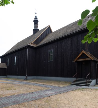 Zdjęcie nr 26: Kościół z drewna modrzewiowego o konstrukcji zrębowej, na betonowej podmurówce, otoczony drewnianym ogrodzeniem. Bryła składa się z korpusu nawowego na planie prostokąta oraz z wydłużonego, prostokątnego prezbiterium z zakrystią na przedłużeniu, węższego od nawy, zakończonego ścianą prostą, skierowanego na południe. Od strony północnej dobudowana jest prostokątna kruchta, od zachodu dwa przedsionki na dwóch skrajnych bokach –  przy nawie i prezbiterium. 
Trójprzęsłowe prezbiterium nakryte jest  sklepieniem pozornym zwierciadlanym, które przedłuża się nad nawę środkową. Ściany prezbiterium artykułowane pilastrami dzielą je na trzy przęsła. Kapitele pilastrów dźwigają nasadniki, które podtrzymują gzymsik. Przedłużeniem kapiteli w strefie stropu są płaskie belki biegnące przez całą jego szerokość, wyznaczając tym samym pola, w których umieszczono polichromię. Po obu stronach ołtarza drzwi w kształcie stojącego prostokąta. Prezbiterium od korpusu nawowego oddzielone silnie wystającą belką sklepienia oraz belką tęczową z grupą Ukrzyżowania. 
Korpus o trzech nawach o układzie halowym, z których środkowa jest dwa razy szersza od bocznych, trójprzęsłowa, z nawami oddzielonymi kanelowanymi filarami z nasadnikami podtrzymującymi gzyms. Nawy boczne nakryte kolebką. Na linii filarów pilastry na ścianach, łączą je poprzeczne belki sklepienia. W ostatnim przęśle chór muzyczny wsparty na dwóch filarach, z pełnym parapetem występującym w części środkowej trójbocznie do przodu. Jego krawędź dolna dekorowana jest lambrekinem. Ściany kościoła pokryte polichormią, pomalowane na jasnożółto, z jasnozielonymi podporami i brązowymi liniami podziału stropu. Te elementy wyznaczają ramy wypełnione polami z dekoracją ornamentalną w kształcie prostokątów i scenami figuralnymi zamkniętymi w kołach. 
Otwory okienne w drugim i trzecim przęśle nawy, od wschodu i zachodu, prostokątne. Także w prezbiterium po jednym prostokątnym oknie z każdej strony. Od strony wejścia dwa niewielkie okna prostokątne ponad kruchtą. Na ścianie zamknięcia zakrystii cztery prostokątne, symetrycznie rozłożone okna. 
 Fasada poprzedzona przedsionkiem w kształcie prostopadłościanu nakrytego dachem dwuspadowym, z przyczółkiem. Otwór drzwiowy w formie stojącego prostokąta, a w szczycie przyczółka trójkątne, wielodzielne okienko. Fasada w kształcie leżącego prostokąta z trójkątnym szczytem, części oddzielone daszkiem pulpitowym. Po dwóch stronach przedsionka prostokątne okna, w szczycie fasady  nakryty daszkiem krucyfiks. 
Elewacje zewnętrzne na podmurówce, artykułowane jedynie pionowymi, wystającymi przed lico belkami, przeprute oknami. Nad nawą i prezbiterium dachy dwuspadowe, nad prezbiterium niższy niż nad nawą, kryty blachą. Nad nawą wieżyczka na sygnaturkę zwieńczona cebulastym hełmem i krzyżem na szczycie. Na końcach dachu przy prezbiterium i nawie metalowe krzyże. 
