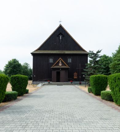 Zdjęcie nr 3: Kościół z drewna modrzewiowego o konstrukcji zrębowej, na betonowej podmurówce, otoczony drewnianym ogrodzeniem. Bryła składa się z korpusu nawowego na planie prostokąta oraz z wydłużonego, prostokątnego prezbiterium z zakrystią na przedłużeniu, węższego od nawy, zakończonego ścianą prostą, skierowanego na południe. Od strony północnej dobudowana jest prostokątna kruchta, od zachodu dwa przedsionki na dwóch skrajnych bokach –  przy nawie i prezbiterium. 
Trójprzęsłowe prezbiterium nakryte jest  sklepieniem pozornym zwierciadlanym, które przedłuża się nad nawę środkową. Ściany prezbiterium artykułowane pilastrami dzielą je na trzy przęsła. Kapitele pilastrów dźwigają nasadniki, które podtrzymują gzymsik. Przedłużeniem kapiteli w strefie stropu są płaskie belki biegnące przez całą jego szerokość, wyznaczając tym samym pola, w których umieszczono polichromię. Po obu stronach ołtarza drzwi w kształcie stojącego prostokąta. Prezbiterium od korpusu nawowego oddzielone silnie wystającą belką sklepienia oraz belką tęczową z grupą Ukrzyżowania. 
Korpus o trzech nawach o układzie halowym, z których środkowa jest dwa razy szersza od bocznych, trójprzęsłowa, z nawami oddzielonymi kanelowanymi filarami z nasadnikami podtrzymującymi gzyms. Nawy boczne nakryte kolebką. Na linii filarów pilastry na ścianach, łączą je poprzeczne belki sklepienia. W ostatnim przęśle chór muzyczny wsparty na dwóch filarach, z pełnym parapetem występującym w części środkowej trójbocznie do przodu. Jego krawędź dolna dekorowana jest lambrekinem. Ściany kościoła pokryte polichormią, pomalowane na jasnożółto, z jasnozielonymi podporami i brązowymi liniami podziału stropu. Te elementy wyznaczają ramy wypełnione polami z dekoracją ornamentalną w kształcie prostokątów i scenami figuralnymi zamkniętymi w kołach. 
Otwory okienne w drugim i trzecim przęśle nawy, od wschodu i zachodu, prostokątne. Także w prezbiterium po jednym prostokątnym oknie z każdej strony. Od strony wejścia dwa niewielkie okna prostokątne ponad kruchtą. Na ścianie zamknięcia zakrystii cztery prostokątne, symetrycznie rozłożone okna. 
 Fasada poprzedzona przedsionkiem w kształcie prostopadłościanu nakrytego dachem dwuspadowym, z przyczółkiem. Otwór drzwiowy w formie stojącego prostokąta, a w szczycie przyczółka trójkątne, wielodzielne okienko. Fasada w kształcie leżącego prostokąta z trójkątnym szczytem, części oddzielone daszkiem pulpitowym. Po dwóch stronach przedsionka prostokątne okna, w szczycie fasady  nakryty daszkiem krucyfiks. 
Elewacje zewnętrzne na podmurówce, artykułowane jedynie pionowymi, wystającymi przed lico belkami, przeprute oknami. Nad nawą i prezbiterium dachy dwuspadowe, nad prezbiterium niższy niż nad nawą, kryty blachą. Nad nawą wieżyczka na sygnaturkę zwieńczona cebulastym hełmem i krzyżem na szczycie. Na końcach dachu przy prezbiterium i nawie metalowe krzyże. 
