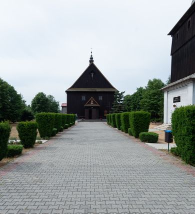 Zdjęcie nr 28: Kościół z drewna modrzewiowego o konstrukcji zrębowej, na betonowej podmurówce, otoczony drewnianym ogrodzeniem. Bryła składa się z korpusu nawowego na planie prostokąta oraz z wydłużonego, prostokątnego prezbiterium z zakrystią na przedłużeniu, węższego od nawy, zakończonego ścianą prostą, skierowanego na południe. Od strony północnej dobudowana jest prostokątna kruchta, od zachodu dwa przedsionki na dwóch skrajnych bokach –  przy nawie i prezbiterium. 
Trójprzęsłowe prezbiterium nakryte jest  sklepieniem pozornym zwierciadlanym, które przedłuża się nad nawę środkową. Ściany prezbiterium artykułowane pilastrami dzielą je na trzy przęsła. Kapitele pilastrów dźwigają nasadniki, które podtrzymują gzymsik. Przedłużeniem kapiteli w strefie stropu są płaskie belki biegnące przez całą jego szerokość, wyznaczając tym samym pola, w których umieszczono polichromię. Po obu stronach ołtarza drzwi w kształcie stojącego prostokąta. Prezbiterium od korpusu nawowego oddzielone silnie wystającą belką sklepienia oraz belką tęczową z grupą Ukrzyżowania. 
Korpus o trzech nawach o układzie halowym, z których środkowa jest dwa razy szersza od bocznych, trójprzęsłowa, z nawami oddzielonymi kanelowanymi filarami z nasadnikami podtrzymującymi gzyms. Nawy boczne nakryte kolebką. Na linii filarów pilastry na ścianach, łączą je poprzeczne belki sklepienia. W ostatnim przęśle chór muzyczny wsparty na dwóch filarach, z pełnym parapetem występującym w części środkowej trójbocznie do przodu. Jego krawędź dolna dekorowana jest lambrekinem. Ściany kościoła pokryte polichormią, pomalowane na jasnożółto, z jasnozielonymi podporami i brązowymi liniami podziału stropu. Te elementy wyznaczają ramy wypełnione polami z dekoracją ornamentalną w kształcie prostokątów i scenami figuralnymi zamkniętymi w kołach. 
Otwory okienne w drugim i trzecim przęśle nawy, od wschodu i zachodu, prostokątne. Także w prezbiterium po jednym prostokątnym oknie z każdej strony. Od strony wejścia dwa niewielkie okna prostokątne ponad kruchtą. Na ścianie zamknięcia zakrystii cztery prostokątne, symetrycznie rozłożone okna. 
 Fasada poprzedzona przedsionkiem w kształcie prostopadłościanu nakrytego dachem dwuspadowym, z przyczółkiem. Otwór drzwiowy w formie stojącego prostokąta, a w szczycie przyczółka trójkątne, wielodzielne okienko. Fasada w kształcie leżącego prostokąta z trójkątnym szczytem, części oddzielone daszkiem pulpitowym. Po dwóch stronach przedsionka prostokątne okna, w szczycie fasady  nakryty daszkiem krucyfiks. 
Elewacje zewnętrzne na podmurówce, artykułowane jedynie pionowymi, wystającymi przed lico belkami, przeprute oknami. Nad nawą i prezbiterium dachy dwuspadowe, nad prezbiterium niższy niż nad nawą, kryty blachą. Nad nawą wieżyczka na sygnaturkę zwieńczona cebulastym hełmem i krzyżem na szczycie. Na końcach dachu przy prezbiterium i nawie metalowe krzyże. 
