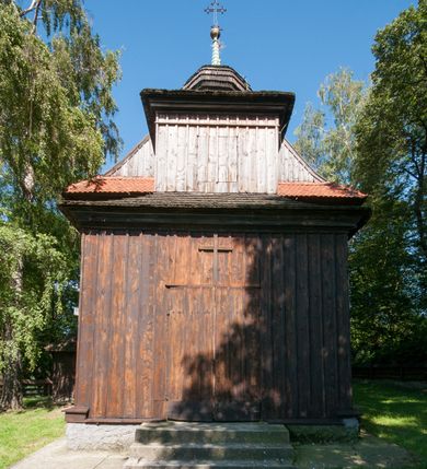 Zdjęcie nr 6: Kościół drewniany, orientowany, jednonawowy, wzniesiony na niewielkiej ceglanej podmurówce, o konstrukcji zrębowej, ściany szalowane pionowo z listowaniem, kruchta zachodnia o konstrukcji słupowo-ramowej. Nawa na rzucie prostokąta zbliżonego do kwadratu, prezbiterium węższe od nawy, wydłużone, zamknięte trójbocznie. Do północnej ściany prezbiterium przylega zakrystia na planie prostokąta. Do północnej ściany nawy przywiera niewielka kruchta na planie prostokąta, zamknięta trójbocznie. Przy zachodniej ścianie nawy znajduje się kruchta na rzucie prostokąta. Wnętrze nakryte pozornymi, spłaszczonymi sklepieniami o krzywiźnie przybierającej kształt łuku odcinkowego. Pomiędzy nawą a prezbiterium otwór tęczowy o wykroju prostokąta zbliżonego do kwadratu zamkniętego łukiem koszowym. Prezbiterium niższe od nawy, sięgające wysokości łuku tęczowego. Ściany prezbiterium i nawy obiega profilowany gzyms, oddzielający je od sklepień. W części zachodniej nawy parapet chóru muzycznego wsparty na dwóch kolumnach o stylizowanych kapitelach kompozytowych i kanelowanych trzonach na wysokich cokołach. W północno-zachodnim narożu drabiniaste schody prowadzące na chór w ośmiobocznej obudowie. Ściany i sklepienia pokryte polichromią. Na ścianach wzdłużnych oraz na osi ściany zachodniej otwory drzwiowe o prostokątnym wykroju zamknięte drzwiami spągowymi z desek oraz płycinowymi, na północnej ścianie prezbiterium prostokątny otwór drzwiowy do zakrystii. Posadzka z płyt kamiennych, szachownicowa. Kruchty i zakrystia nakryte stropami, przez nie dostęp do wnętrza. 
Elewacja frontowa jednoosiowa, zamknięta trójkątnym szczytem, poprzedzona kruchtą. Na osi wejście do kruchty zachodniej w postaci dwuskrzydłowych drzwi w formie stojącego prostokąta. Nad kruchtą pięterko dzwonowe z prostokątnym otworem zamykanym dwuskrzydłowymi okiennicami. Elewacja boczna południowa z czterema oknami, po dwa w nawie i prezbiterium. Elewacja północna przepruta dwoma oknami w części nawy. Okna krosnowe, w formie stojącego prostokąta, o niewielkich podziałach kwaterowych z witrażami. Na wschodniej ścianie kruchty północnej okno i jednoskrzydłowe drzwi. W zakrystii okno na ścianie północnej i drzwi jednoskrzydłowe na ścianie wschodniej. Kościół nakrywają dachy dwuspadowe, wyższy nad korpusem nawowym, niższy nad prezbiterium. Kruchta zachodnia w formie przysadzistej wieży, niższej od korpusu nawowego z pięterkiem dzwonowym, zwieńczonym hełmem baniastym, pokrytym gontem z iglicą oraz gałką i krzyżem. Kruchta północna z dachem wielospadowym. Zakrystia nakryta dachem pulpitowym. W połowie wysokości dachu znajduje się wieżyczka na sygnaturkę na ośmiobocznym obitym blachą trzonie, z ażurową latarnią nakrytą baniastym hełmem zakończonym kulą z krzyżem. 



