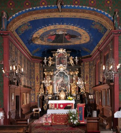 Zdjęcie nr 8: Kościół drewniany, orientowany, jednonawowy, wzniesiony na niewielkiej ceglanej podmurówce, o konstrukcji zrębowej, ściany szalowane pionowo z listowaniem, kruchta zachodnia o konstrukcji słupowo-ramowej. Nawa na rzucie prostokąta zbliżonego do kwadratu, prezbiterium węższe od nawy, wydłużone, zamknięte trójbocznie. Do północnej ściany prezbiterium przylega zakrystia na planie prostokąta. Do północnej ściany nawy przywiera niewielka kruchta na planie prostokąta, zamknięta trójbocznie. Przy zachodniej ścianie nawy znajduje się kruchta na rzucie prostokąta. Wnętrze nakryte pozornymi, spłaszczonymi sklepieniami o krzywiźnie przybierającej kształt łuku odcinkowego. Pomiędzy nawą a prezbiterium otwór tęczowy o wykroju prostokąta zbliżonego do kwadratu zamkniętego łukiem koszowym. Prezbiterium niższe od nawy, sięgające wysokości łuku tęczowego. Ściany prezbiterium i nawy obiega profilowany gzyms, oddzielający je od sklepień. W części zachodniej nawy parapet chóru muzycznego wsparty na dwóch kolumnach o stylizowanych kapitelach kompozytowych i kanelowanych trzonach na wysokich cokołach. W północno-zachodnim narożu drabiniaste schody prowadzące na chór w ośmiobocznej obudowie. Ściany i sklepienia pokryte polichromią. Na ścianach wzdłużnych oraz na osi ściany zachodniej otwory drzwiowe o prostokątnym wykroju zamknięte drzwiami spągowymi z desek oraz płycinowymi, na północnej ścianie prezbiterium prostokątny otwór drzwiowy do zakrystii. Posadzka z płyt kamiennych, szachownicowa. Kruchty i zakrystia nakryte stropami, przez nie dostęp do wnętrza. 
Elewacja frontowa jednoosiowa, zamknięta trójkątnym szczytem, poprzedzona kruchtą. Na osi wejście do kruchty zachodniej w postaci dwuskrzydłowych drzwi w formie stojącego prostokąta. Nad kruchtą pięterko dzwonowe z prostokątnym otworem zamykanym dwuskrzydłowymi okiennicami. Elewacja boczna południowa z czterema oknami, po dwa w nawie i prezbiterium. Elewacja północna przepruta dwoma oknami w części nawy. Okna krosnowe, w formie stojącego prostokąta, o niewielkich podziałach kwaterowych z witrażami. Na wschodniej ścianie kruchty północnej okno i jednoskrzydłowe drzwi. W zakrystii okno na ścianie północnej i drzwi jednoskrzydłowe na ścianie wschodniej. Kościół nakrywają dachy dwuspadowe, wyższy nad korpusem nawowym, niższy nad prezbiterium. Kruchta zachodnia w formie przysadzistej wieży, niższej od korpusu nawowego z pięterkiem dzwonowym, zwieńczonym hełmem baniastym, pokrytym gontem z iglicą oraz gałką i krzyżem. Kruchta północna z dachem wielospadowym. Zakrystia nakryta dachem pulpitowym. W połowie wysokości dachu znajduje się wieżyczka na sygnaturkę na ośmiobocznym obitym blachą trzonie, z ażurową latarnią nakrytą baniastym hełmem zakończonym kulą z krzyżem. 



