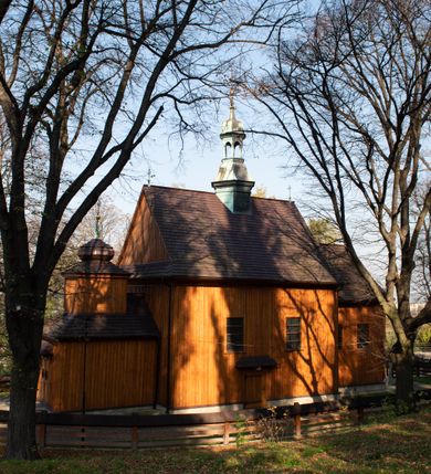 Zdjęcie nr 20: Kościół drewniany, orientowany, jednonawowy, wzniesiony na niewielkiej ceglanej podmurówce, o konstrukcji zrębowej, ściany szalowane pionowo z listowaniem, kruchta zachodnia o konstrukcji słupowo-ramowej. Nawa na rzucie prostokąta zbliżonego do kwadratu, prezbiterium węższe od nawy, wydłużone, zamknięte trójbocznie. Do północnej ściany prezbiterium przylega zakrystia na planie prostokąta. Do północnej ściany nawy przywiera niewielka kruchta na planie prostokąta, zamknięta trójbocznie. Przy zachodniej ścianie nawy znajduje się kruchta na rzucie prostokąta. Wnętrze nakryte pozornymi, spłaszczonymi sklepieniami o krzywiźnie przybierającej kształt łuku odcinkowego. Pomiędzy nawą a prezbiterium otwór tęczowy o wykroju prostokąta zbliżonego do kwadratu zamkniętego łukiem koszowym. Prezbiterium niższe od nawy, sięgające wysokości łuku tęczowego. Ściany prezbiterium i nawy obiega profilowany gzyms, oddzielający je od sklepień. W części zachodniej nawy parapet chóru muzycznego wsparty na dwóch kolumnach o stylizowanych kapitelach kompozytowych i kanelowanych trzonach na wysokich cokołach. W północno-zachodnim narożu drabiniaste schody prowadzące na chór w ośmiobocznej obudowie. Ściany i sklepienia pokryte polichromią. Na ścianach wzdłużnych oraz na osi ściany zachodniej otwory drzwiowe o prostokątnym wykroju zamknięte drzwiami spągowymi z desek oraz płycinowymi, na północnej ścianie prezbiterium prostokątny otwór drzwiowy do zakrystii. Posadzka z płyt kamiennych, szachownicowa. Kruchty i zakrystia nakryte stropami, przez nie dostęp do wnętrza. 
Elewacja frontowa jednoosiowa, zamknięta trójkątnym szczytem, poprzedzona kruchtą. Na osi wejście do kruchty zachodniej w postaci dwuskrzydłowych drzwi w formie stojącego prostokąta. Nad kruchtą pięterko dzwonowe z prostokątnym otworem zamykanym dwuskrzydłowymi okiennicami. Elewacja boczna południowa z czterema oknami, po dwa w nawie i prezbiterium. Elewacja północna przepruta dwoma oknami w części nawy. Okna krosnowe, w formie stojącego prostokąta, o niewielkich podziałach kwaterowych z witrażami. Na wschodniej ścianie kruchty północnej okno i jednoskrzydłowe drzwi. W zakrystii okno na ścianie północnej i drzwi jednoskrzydłowe na ścianie wschodniej. Kościół nakrywają dachy dwuspadowe, wyższy nad korpusem nawowym, niższy nad prezbiterium. Kruchta zachodnia w formie przysadzistej wieży, niższej od korpusu nawowego z pięterkiem dzwonowym, zwieńczonym hełmem baniastym, pokrytym gontem z iglicą oraz gałką i krzyżem. Kruchta północna z dachem wielospadowym. Zakrystia nakryta dachem pulpitowym. W połowie wysokości dachu znajduje się wieżyczka na sygnaturkę na ośmiobocznym obitym blachą trzonie, z ażurową latarnią nakrytą baniastym hełmem zakończonym kulą z krzyżem. 



