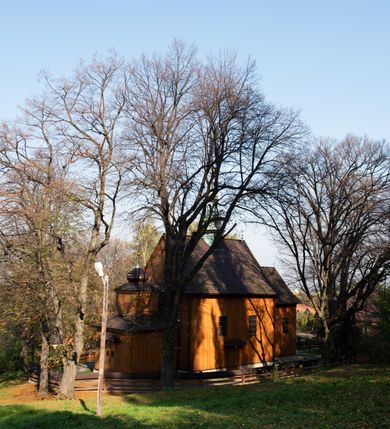 Zdjęcie nr 22: Kościół drewniany, orientowany, jednonawowy, wzniesiony na niewielkiej ceglanej podmurówce, o konstrukcji zrębowej, ściany szalowane pionowo z listowaniem, kruchta zachodnia o konstrukcji słupowo-ramowej. Nawa na rzucie prostokąta zbliżonego do kwadratu, prezbiterium węższe od nawy, wydłużone, zamknięte trójbocznie. Do północnej ściany prezbiterium przylega zakrystia na planie prostokąta. Do północnej ściany nawy przywiera niewielka kruchta na planie prostokąta, zamknięta trójbocznie. Przy zachodniej ścianie nawy znajduje się kruchta na rzucie prostokąta. Wnętrze nakryte pozornymi, spłaszczonymi sklepieniami o krzywiźnie przybierającej kształt łuku odcinkowego. Pomiędzy nawą a prezbiterium otwór tęczowy o wykroju prostokąta zbliżonego do kwadratu zamkniętego łukiem koszowym. Prezbiterium niższe od nawy, sięgające wysokości łuku tęczowego. Ściany prezbiterium i nawy obiega profilowany gzyms, oddzielający je od sklepień. W części zachodniej nawy parapet chóru muzycznego wsparty na dwóch kolumnach o stylizowanych kapitelach kompozytowych i kanelowanych trzonach na wysokich cokołach. W północno-zachodnim narożu drabiniaste schody prowadzące na chór w ośmiobocznej obudowie. Ściany i sklepienia pokryte polichromią. Na ścianach wzdłużnych oraz na osi ściany zachodniej otwory drzwiowe o prostokątnym wykroju zamknięte drzwiami spągowymi z desek oraz płycinowymi, na północnej ścianie prezbiterium prostokątny otwór drzwiowy do zakrystii. Posadzka z płyt kamiennych, szachownicowa. Kruchty i zakrystia nakryte stropami, przez nie dostęp do wnętrza. 
Elewacja frontowa jednoosiowa, zamknięta trójkątnym szczytem, poprzedzona kruchtą. Na osi wejście do kruchty zachodniej w postaci dwuskrzydłowych drzwi w formie stojącego prostokąta. Nad kruchtą pięterko dzwonowe z prostokątnym otworem zamykanym dwuskrzydłowymi okiennicami. Elewacja boczna południowa z czterema oknami, po dwa w nawie i prezbiterium. Elewacja północna przepruta dwoma oknami w części nawy. Okna krosnowe, w formie stojącego prostokąta, o niewielkich podziałach kwaterowych z witrażami. Na wschodniej ścianie kruchty północnej okno i jednoskrzydłowe drzwi. W zakrystii okno na ścianie północnej i drzwi jednoskrzydłowe na ścianie wschodniej. Kościół nakrywają dachy dwuspadowe, wyższy nad korpusem nawowym, niższy nad prezbiterium. Kruchta zachodnia w formie przysadzistej wieży, niższej od korpusu nawowego z pięterkiem dzwonowym, zwieńczonym hełmem baniastym, pokrytym gontem z iglicą oraz gałką i krzyżem. Kruchta północna z dachem wielospadowym. Zakrystia nakryta dachem pulpitowym. W połowie wysokości dachu znajduje się wieżyczka na sygnaturkę na ośmiobocznym obitym blachą trzonie, z ażurową latarnią nakrytą baniastym hełmem zakończonym kulą z krzyżem. 



