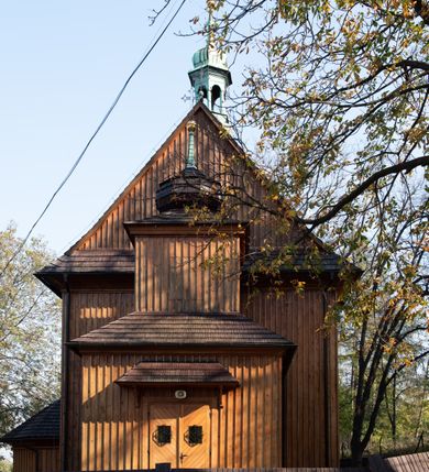 Zdjęcie nr 26: Kościół drewniany, orientowany, jednonawowy, wzniesiony na niewielkiej ceglanej podmurówce, o konstrukcji zrębowej, ściany szalowane pionowo z listowaniem, kruchta zachodnia o konstrukcji słupowo-ramowej. Nawa na rzucie prostokąta zbliżonego do kwadratu, prezbiterium węższe od nawy, wydłużone, zamknięte trójbocznie. Do północnej ściany prezbiterium przylega zakrystia na planie prostokąta. Do północnej ściany nawy przywiera niewielka kruchta na planie prostokąta, zamknięta trójbocznie. Przy zachodniej ścianie nawy znajduje się kruchta na rzucie prostokąta. Wnętrze nakryte pozornymi, spłaszczonymi sklepieniami o krzywiźnie przybierającej kształt łuku odcinkowego. Pomiędzy nawą a prezbiterium otwór tęczowy o wykroju prostokąta zbliżonego do kwadratu zamkniętego łukiem koszowym. Prezbiterium niższe od nawy, sięgające wysokości łuku tęczowego. Ściany prezbiterium i nawy obiega profilowany gzyms, oddzielający je od sklepień. W części zachodniej nawy parapet chóru muzycznego wsparty na dwóch kolumnach o stylizowanych kapitelach kompozytowych i kanelowanych trzonach na wysokich cokołach. W północno-zachodnim narożu drabiniaste schody prowadzące na chór w ośmiobocznej obudowie. Ściany i sklepienia pokryte polichromią. Na ścianach wzdłużnych oraz na osi ściany zachodniej otwory drzwiowe o prostokątnym wykroju zamknięte drzwiami spągowymi z desek oraz płycinowymi, na północnej ścianie prezbiterium prostokątny otwór drzwiowy do zakrystii. Posadzka z płyt kamiennych, szachownicowa. Kruchty i zakrystia nakryte stropami, przez nie dostęp do wnętrza. 
Elewacja frontowa jednoosiowa, zamknięta trójkątnym szczytem, poprzedzona kruchtą. Na osi wejście do kruchty zachodniej w postaci dwuskrzydłowych drzwi w formie stojącego prostokąta. Nad kruchtą pięterko dzwonowe z prostokątnym otworem zamykanym dwuskrzydłowymi okiennicami. Elewacja boczna południowa z czterema oknami, po dwa w nawie i prezbiterium. Elewacja północna przepruta dwoma oknami w części nawy. Okna krosnowe, w formie stojącego prostokąta, o niewielkich podziałach kwaterowych z witrażami. Na wschodniej ścianie kruchty północnej okno i jednoskrzydłowe drzwi. W zakrystii okno na ścianie północnej i drzwi jednoskrzydłowe na ścianie wschodniej. Kościół nakrywają dachy dwuspadowe, wyższy nad korpusem nawowym, niższy nad prezbiterium. Kruchta zachodnia w formie przysadzistej wieży, niższej od korpusu nawowego z pięterkiem dzwonowym, zwieńczonym hełmem baniastym, pokrytym gontem z iglicą oraz gałką i krzyżem. Kruchta północna z dachem wielospadowym. Zakrystia nakryta dachem pulpitowym. W połowie wysokości dachu znajduje się wieżyczka na sygnaturkę na ośmiobocznym obitym blachą trzonie, z ażurową latarnią nakrytą baniastym hełmem zakończonym kulą z krzyżem. 



