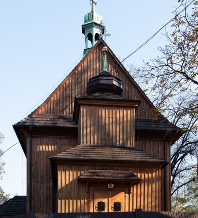 Zdjęcie nr 25: Kościół drewniany, orientowany, jednonawowy, wzniesiony na niewielkiej ceglanej podmurówce, o konstrukcji zrębowej, ściany szalowane pionowo z listowaniem, kruchta zachodnia o konstrukcji słupowo-ramowej. Nawa na rzucie prostokąta zbliżonego do kwadratu, prezbiterium węższe od nawy, wydłużone, zamknięte trójbocznie. Do północnej ściany prezbiterium przylega zakrystia na planie prostokąta. Do północnej ściany nawy przywiera niewielka kruchta na planie prostokąta, zamknięta trójbocznie. Przy zachodniej ścianie nawy znajduje się kruchta na rzucie prostokąta. Wnętrze nakryte pozornymi, spłaszczonymi sklepieniami o krzywiźnie przybierającej kształt łuku odcinkowego. Pomiędzy nawą a prezbiterium otwór tęczowy o wykroju prostokąta zbliżonego do kwadratu zamkniętego łukiem koszowym. Prezbiterium niższe od nawy, sięgające wysokości łuku tęczowego. Ściany prezbiterium i nawy obiega profilowany gzyms, oddzielający je od sklepień. W części zachodniej nawy parapet chóru muzycznego wsparty na dwóch kolumnach o stylizowanych kapitelach kompozytowych i kanelowanych trzonach na wysokich cokołach. W północno-zachodnim narożu drabiniaste schody prowadzące na chór w ośmiobocznej obudowie. Ściany i sklepienia pokryte polichromią. Na ścianach wzdłużnych oraz na osi ściany zachodniej otwory drzwiowe o prostokątnym wykroju zamknięte drzwiami spągowymi z desek oraz płycinowymi, na północnej ścianie prezbiterium prostokątny otwór drzwiowy do zakrystii. Posadzka z płyt kamiennych, szachownicowa. Kruchty i zakrystia nakryte stropami, przez nie dostęp do wnętrza. 
Elewacja frontowa jednoosiowa, zamknięta trójkątnym szczytem, poprzedzona kruchtą. Na osi wejście do kruchty zachodniej w postaci dwuskrzydłowych drzwi w formie stojącego prostokąta. Nad kruchtą pięterko dzwonowe z prostokątnym otworem zamykanym dwuskrzydłowymi okiennicami. Elewacja boczna południowa z czterema oknami, po dwa w nawie i prezbiterium. Elewacja północna przepruta dwoma oknami w części nawy. Okna krosnowe, w formie stojącego prostokąta, o niewielkich podziałach kwaterowych z witrażami. Na wschodniej ścianie kruchty północnej okno i jednoskrzydłowe drzwi. W zakrystii okno na ścianie północnej i drzwi jednoskrzydłowe na ścianie wschodniej. Kościół nakrywają dachy dwuspadowe, wyższy nad korpusem nawowym, niższy nad prezbiterium. Kruchta zachodnia w formie przysadzistej wieży, niższej od korpusu nawowego z pięterkiem dzwonowym, zwieńczonym hełmem baniastym, pokrytym gontem z iglicą oraz gałką i krzyżem. Kruchta północna z dachem wielospadowym. Zakrystia nakryta dachem pulpitowym. W połowie wysokości dachu znajduje się wieżyczka na sygnaturkę na ośmiobocznym obitym blachą trzonie, z ażurową latarnią nakrytą baniastym hełmem zakończonym kulą z krzyżem. 



