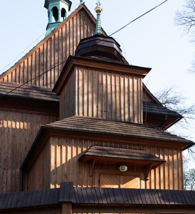 Zdjęcie nr 28: Kościół drewniany, orientowany, jednonawowy, wzniesiony na niewielkiej ceglanej podmurówce, o konstrukcji zrębowej, ściany szalowane pionowo z listowaniem, kruchta zachodnia o konstrukcji słupowo-ramowej. Nawa na rzucie prostokąta zbliżonego do kwadratu, prezbiterium węższe od nawy, wydłużone, zamknięte trójbocznie. Do północnej ściany prezbiterium przylega zakrystia na planie prostokąta. Do północnej ściany nawy przywiera niewielka kruchta na planie prostokąta, zamknięta trójbocznie. Przy zachodniej ścianie nawy znajduje się kruchta na rzucie prostokąta. Wnętrze nakryte pozornymi, spłaszczonymi sklepieniami o krzywiźnie przybierającej kształt łuku odcinkowego. Pomiędzy nawą a prezbiterium otwór tęczowy o wykroju prostokąta zbliżonego do kwadratu zamkniętego łukiem koszowym. Prezbiterium niższe od nawy, sięgające wysokości łuku tęczowego. Ściany prezbiterium i nawy obiega profilowany gzyms, oddzielający je od sklepień. W części zachodniej nawy parapet chóru muzycznego wsparty na dwóch kolumnach o stylizowanych kapitelach kompozytowych i kanelowanych trzonach na wysokich cokołach. W północno-zachodnim narożu drabiniaste schody prowadzące na chór w ośmiobocznej obudowie. Ściany i sklepienia pokryte polichromią. Na ścianach wzdłużnych oraz na osi ściany zachodniej otwory drzwiowe o prostokątnym wykroju zamknięte drzwiami spągowymi z desek oraz płycinowymi, na północnej ścianie prezbiterium prostokątny otwór drzwiowy do zakrystii. Posadzka z płyt kamiennych, szachownicowa. Kruchty i zakrystia nakryte stropami, przez nie dostęp do wnętrza. 
Elewacja frontowa jednoosiowa, zamknięta trójkątnym szczytem, poprzedzona kruchtą. Na osi wejście do kruchty zachodniej w postaci dwuskrzydłowych drzwi w formie stojącego prostokąta. Nad kruchtą pięterko dzwonowe z prostokątnym otworem zamykanym dwuskrzydłowymi okiennicami. Elewacja boczna południowa z czterema oknami, po dwa w nawie i prezbiterium. Elewacja północna przepruta dwoma oknami w części nawy. Okna krosnowe, w formie stojącego prostokąta, o niewielkich podziałach kwaterowych z witrażami. Na wschodniej ścianie kruchty północnej okno i jednoskrzydłowe drzwi. W zakrystii okno na ścianie północnej i drzwi jednoskrzydłowe na ścianie wschodniej. Kościół nakrywają dachy dwuspadowe, wyższy nad korpusem nawowym, niższy nad prezbiterium. Kruchta zachodnia w formie przysadzistej wieży, niższej od korpusu nawowego z pięterkiem dzwonowym, zwieńczonym hełmem baniastym, pokrytym gontem z iglicą oraz gałką i krzyżem. Kruchta północna z dachem wielospadowym. Zakrystia nakryta dachem pulpitowym. W połowie wysokości dachu znajduje się wieżyczka na sygnaturkę na ośmiobocznym obitym blachą trzonie, z ażurową latarnią nakrytą baniastym hełmem zakończonym kulą z krzyżem. 



