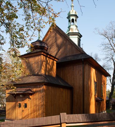 Zdjęcie nr 29: Kościół drewniany, orientowany, jednonawowy, wzniesiony na niewielkiej ceglanej podmurówce, o konstrukcji zrębowej, ściany szalowane pionowo z listowaniem, kruchta zachodnia o konstrukcji słupowo-ramowej. Nawa na rzucie prostokąta zbliżonego do kwadratu, prezbiterium węższe od nawy, wydłużone, zamknięte trójbocznie. Do północnej ściany prezbiterium przylega zakrystia na planie prostokąta. Do północnej ściany nawy przywiera niewielka kruchta na planie prostokąta, zamknięta trójbocznie. Przy zachodniej ścianie nawy znajduje się kruchta na rzucie prostokąta. Wnętrze nakryte pozornymi, spłaszczonymi sklepieniami o krzywiźnie przybierającej kształt łuku odcinkowego. Pomiędzy nawą a prezbiterium otwór tęczowy o wykroju prostokąta zbliżonego do kwadratu zamkniętego łukiem koszowym. Prezbiterium niższe od nawy, sięgające wysokości łuku tęczowego. Ściany prezbiterium i nawy obiega profilowany gzyms, oddzielający je od sklepień. W części zachodniej nawy parapet chóru muzycznego wsparty na dwóch kolumnach o stylizowanych kapitelach kompozytowych i kanelowanych trzonach na wysokich cokołach. W północno-zachodnim narożu drabiniaste schody prowadzące na chór w ośmiobocznej obudowie. Ściany i sklepienia pokryte polichromią. Na ścianach wzdłużnych oraz na osi ściany zachodniej otwory drzwiowe o prostokątnym wykroju zamknięte drzwiami spągowymi z desek oraz płycinowymi, na północnej ścianie prezbiterium prostokątny otwór drzwiowy do zakrystii. Posadzka z płyt kamiennych, szachownicowa. Kruchty i zakrystia nakryte stropami, przez nie dostęp do wnętrza. 
Elewacja frontowa jednoosiowa, zamknięta trójkątnym szczytem, poprzedzona kruchtą. Na osi wejście do kruchty zachodniej w postaci dwuskrzydłowych drzwi w formie stojącego prostokąta. Nad kruchtą pięterko dzwonowe z prostokątnym otworem zamykanym dwuskrzydłowymi okiennicami. Elewacja boczna południowa z czterema oknami, po dwa w nawie i prezbiterium. Elewacja północna przepruta dwoma oknami w części nawy. Okna krosnowe, w formie stojącego prostokąta, o niewielkich podziałach kwaterowych z witrażami. Na wschodniej ścianie kruchty północnej okno i jednoskrzydłowe drzwi. W zakrystii okno na ścianie północnej i drzwi jednoskrzydłowe na ścianie wschodniej. Kościół nakrywają dachy dwuspadowe, wyższy nad korpusem nawowym, niższy nad prezbiterium. Kruchta zachodnia w formie przysadzistej wieży, niższej od korpusu nawowego z pięterkiem dzwonowym, zwieńczonym hełmem baniastym, pokrytym gontem z iglicą oraz gałką i krzyżem. Kruchta północna z dachem wielospadowym. Zakrystia nakryta dachem pulpitowym. W połowie wysokości dachu znajduje się wieżyczka na sygnaturkę na ośmiobocznym obitym blachą trzonie, z ażurową latarnią nakrytą baniastym hełmem zakończonym kulą z krzyżem. 



