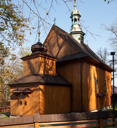 Zdjęcie nr 30: Kościół drewniany, orientowany, jednonawowy, wzniesiony na niewielkiej ceglanej podmurówce, o konstrukcji zrębowej, ściany szalowane pionowo z listowaniem, kruchta zachodnia o konstrukcji słupowo-ramowej. Nawa na rzucie prostokąta zbliżonego do kwadratu, prezbiterium węższe od nawy, wydłużone, zamknięte trójbocznie. Do północnej ściany prezbiterium przylega zakrystia na planie prostokąta. Do północnej ściany nawy przywiera niewielka kruchta na planie prostokąta, zamknięta trójbocznie. Przy zachodniej ścianie nawy znajduje się kruchta na rzucie prostokąta. Wnętrze nakryte pozornymi, spłaszczonymi sklepieniami o krzywiźnie przybierającej kształt łuku odcinkowego. Pomiędzy nawą a prezbiterium otwór tęczowy o wykroju prostokąta zbliżonego do kwadratu zamkniętego łukiem koszowym. Prezbiterium niższe od nawy, sięgające wysokości łuku tęczowego. Ściany prezbiterium i nawy obiega profilowany gzyms, oddzielający je od sklepień. W części zachodniej nawy parapet chóru muzycznego wsparty na dwóch kolumnach o stylizowanych kapitelach kompozytowych i kanelowanych trzonach na wysokich cokołach. W północno-zachodnim narożu drabiniaste schody prowadzące na chór w ośmiobocznej obudowie. Ściany i sklepienia pokryte polichromią. Na ścianach wzdłużnych oraz na osi ściany zachodniej otwory drzwiowe o prostokątnym wykroju zamknięte drzwiami spągowymi z desek oraz płycinowymi, na północnej ścianie prezbiterium prostokątny otwór drzwiowy do zakrystii. Posadzka z płyt kamiennych, szachownicowa. Kruchty i zakrystia nakryte stropami, przez nie dostęp do wnętrza. 
Elewacja frontowa jednoosiowa, zamknięta trójkątnym szczytem, poprzedzona kruchtą. Na osi wejście do kruchty zachodniej w postaci dwuskrzydłowych drzwi w formie stojącego prostokąta. Nad kruchtą pięterko dzwonowe z prostokątnym otworem zamykanym dwuskrzydłowymi okiennicami. Elewacja boczna południowa z czterema oknami, po dwa w nawie i prezbiterium. Elewacja północna przepruta dwoma oknami w części nawy. Okna krosnowe, w formie stojącego prostokąta, o niewielkich podziałach kwaterowych z witrażami. Na wschodniej ścianie kruchty północnej okno i jednoskrzydłowe drzwi. W zakrystii okno na ścianie północnej i drzwi jednoskrzydłowe na ścianie wschodniej. Kościół nakrywają dachy dwuspadowe, wyższy nad korpusem nawowym, niższy nad prezbiterium. Kruchta zachodnia w formie przysadzistej wieży, niższej od korpusu nawowego z pięterkiem dzwonowym, zwieńczonym hełmem baniastym, pokrytym gontem z iglicą oraz gałką i krzyżem. Kruchta północna z dachem wielospadowym. Zakrystia nakryta dachem pulpitowym. W połowie wysokości dachu znajduje się wieżyczka na sygnaturkę na ośmiobocznym obitym blachą trzonie, z ażurową latarnią nakrytą baniastym hełmem zakończonym kulą z krzyżem. 



