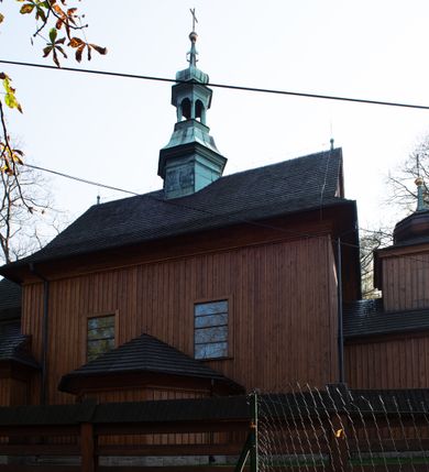 Zdjęcie nr 37: Kościół drewniany, orientowany, jednonawowy, wzniesiony na niewielkiej ceglanej podmurówce, o konstrukcji zrębowej, ściany szalowane pionowo z listowaniem, kruchta zachodnia o konstrukcji słupowo-ramowej. Nawa na rzucie prostokąta zbliżonego do kwadratu, prezbiterium węższe od nawy, wydłużone, zamknięte trójbocznie. Do północnej ściany prezbiterium przylega zakrystia na planie prostokąta. Do północnej ściany nawy przywiera niewielka kruchta na planie prostokąta, zamknięta trójbocznie. Przy zachodniej ścianie nawy znajduje się kruchta na rzucie prostokąta. Wnętrze nakryte pozornymi, spłaszczonymi sklepieniami o krzywiźnie przybierającej kształt łuku odcinkowego. Pomiędzy nawą a prezbiterium otwór tęczowy o wykroju prostokąta zbliżonego do kwadratu zamkniętego łukiem koszowym. Prezbiterium niższe od nawy, sięgające wysokości łuku tęczowego. Ściany prezbiterium i nawy obiega profilowany gzyms, oddzielający je od sklepień. W części zachodniej nawy parapet chóru muzycznego wsparty na dwóch kolumnach o stylizowanych kapitelach kompozytowych i kanelowanych trzonach na wysokich cokołach. W północno-zachodnim narożu drabiniaste schody prowadzące na chór w ośmiobocznej obudowie. Ściany i sklepienia pokryte polichromią. Na ścianach wzdłużnych oraz na osi ściany zachodniej otwory drzwiowe o prostokątnym wykroju zamknięte drzwiami spągowymi z desek oraz płycinowymi, na północnej ścianie prezbiterium prostokątny otwór drzwiowy do zakrystii. Posadzka z płyt kamiennych, szachownicowa. Kruchty i zakrystia nakryte stropami, przez nie dostęp do wnętrza. 
Elewacja frontowa jednoosiowa, zamknięta trójkątnym szczytem, poprzedzona kruchtą. Na osi wejście do kruchty zachodniej w postaci dwuskrzydłowych drzwi w formie stojącego prostokąta. Nad kruchtą pięterko dzwonowe z prostokątnym otworem zamykanym dwuskrzydłowymi okiennicami. Elewacja boczna południowa z czterema oknami, po dwa w nawie i prezbiterium. Elewacja północna przepruta dwoma oknami w części nawy. Okna krosnowe, w formie stojącego prostokąta, o niewielkich podziałach kwaterowych z witrażami. Na wschodniej ścianie kruchty północnej okno i jednoskrzydłowe drzwi. W zakrystii okno na ścianie północnej i drzwi jednoskrzydłowe na ścianie wschodniej. Kościół nakrywają dachy dwuspadowe, wyższy nad korpusem nawowym, niższy nad prezbiterium. Kruchta zachodnia w formie przysadzistej wieży, niższej od korpusu nawowego z pięterkiem dzwonowym, zwieńczonym hełmem baniastym, pokrytym gontem z iglicą oraz gałką i krzyżem. Kruchta północna z dachem wielospadowym. Zakrystia nakryta dachem pulpitowym. W połowie wysokości dachu znajduje się wieżyczka na sygnaturkę na ośmiobocznym obitym blachą trzonie, z ażurową latarnią nakrytą baniastym hełmem zakończonym kulą z krzyżem. 



